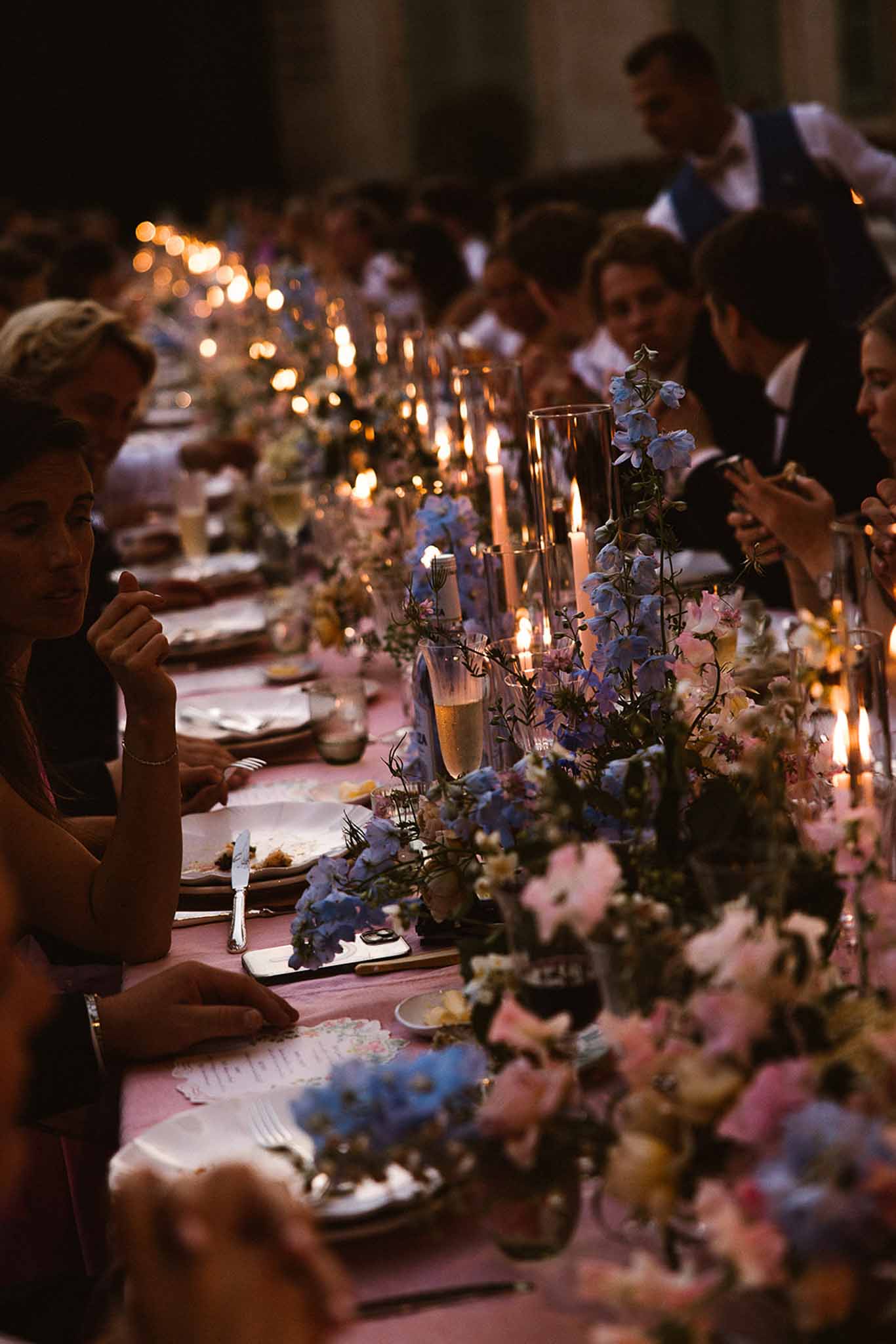Candlelit long table with blue delphinium and pink sweet pea runner on dusty rose linen at evening dinner