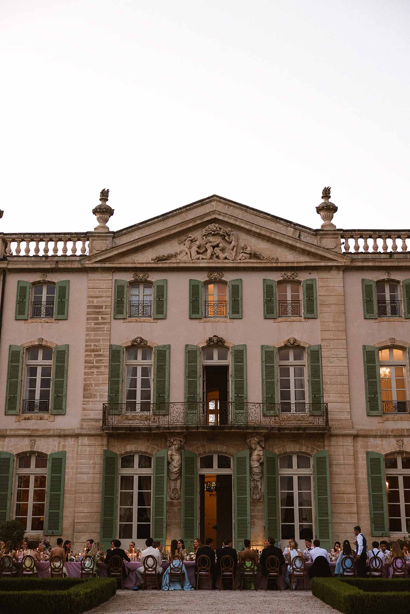 Outdoor reception dinner with guests seated at long tables in front of a pink-rendered French chateau at dusk