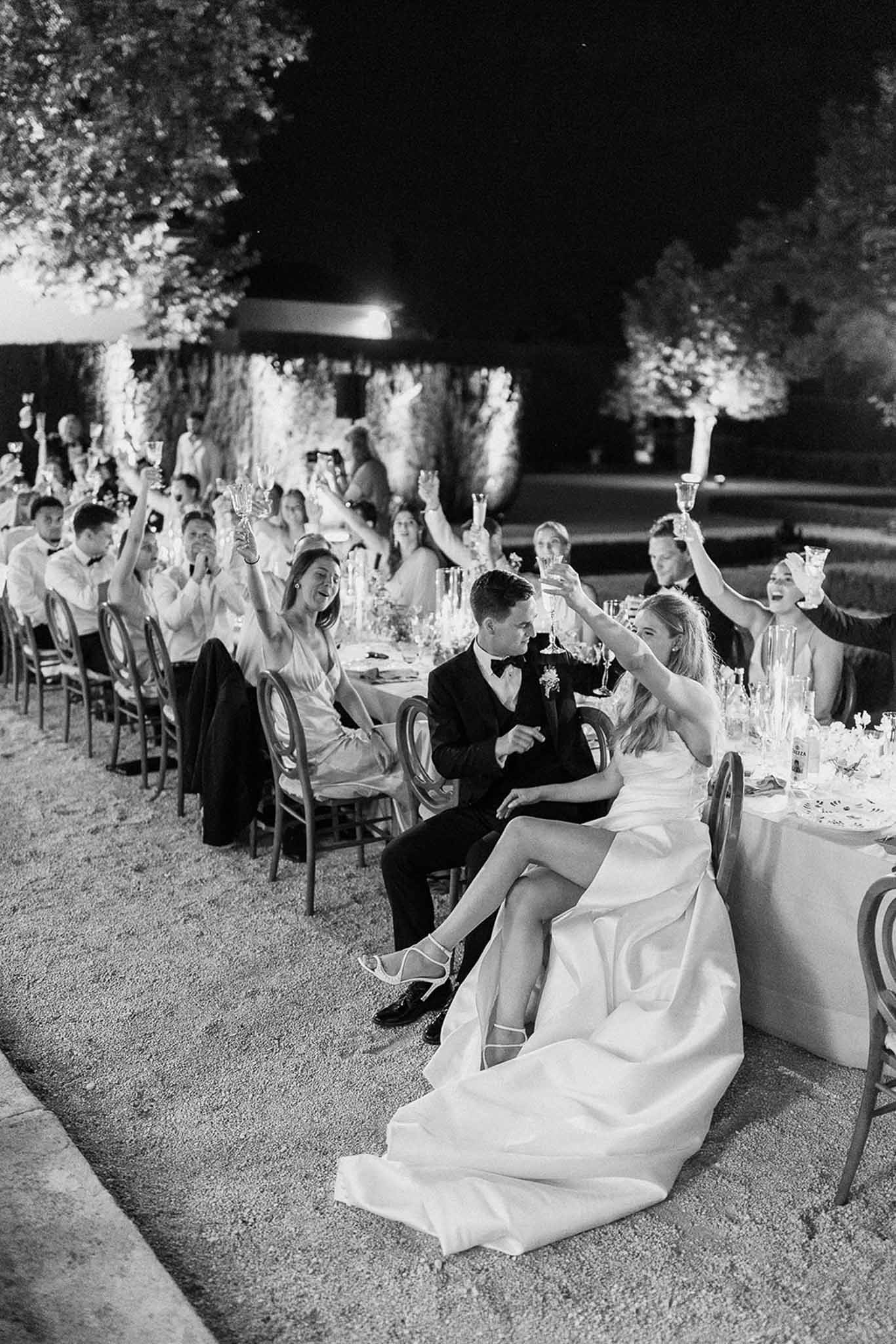 Champagne toast at long reception table with 20-plus guests on outdoor terrace at night in B&W