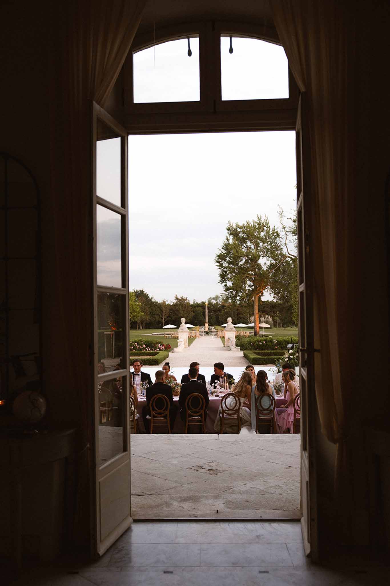 Wedding reception dinner viewed through chateau arched doors onto terrace with formal French garden beyond