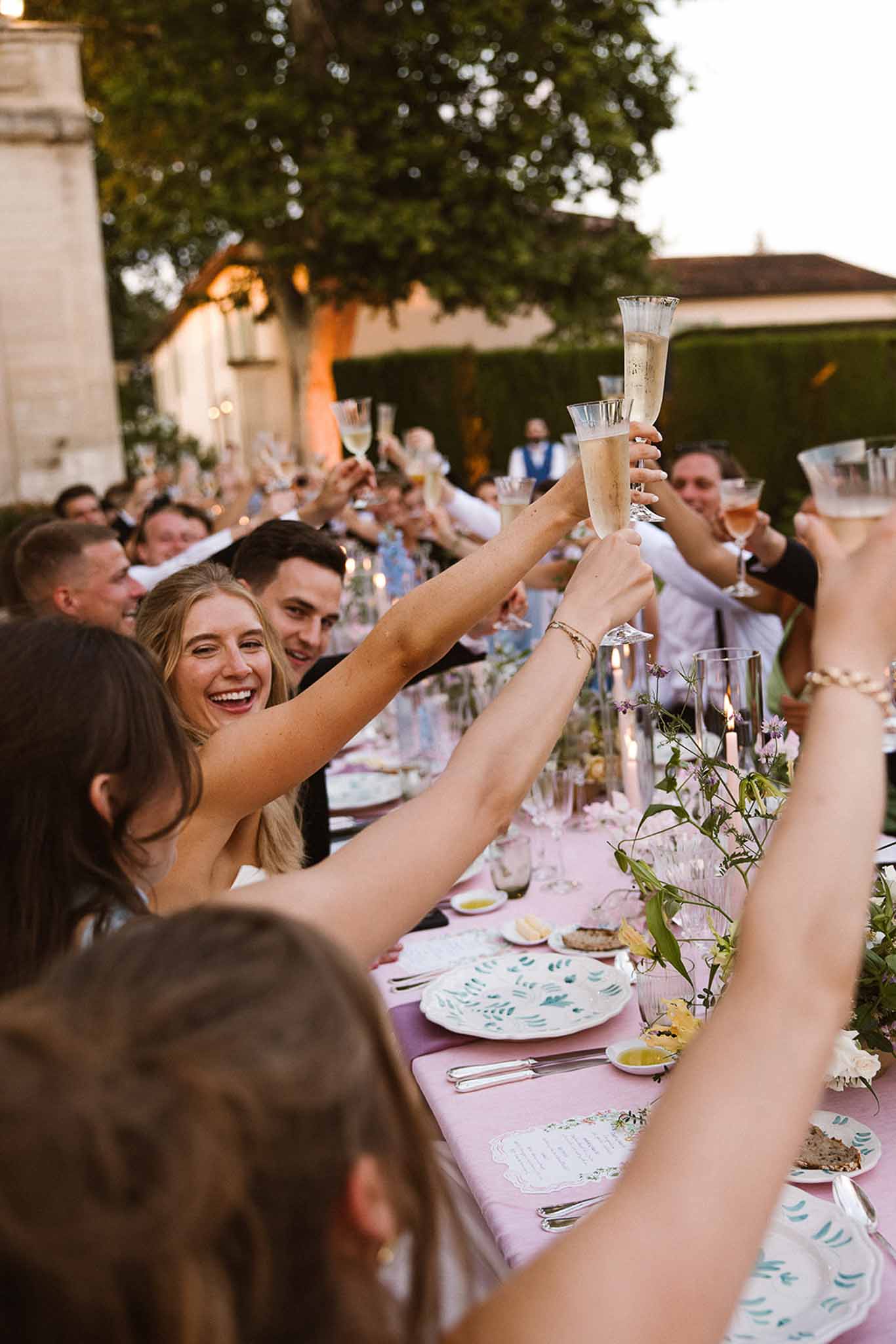 Guests raising glasses in toast at long outdoor reception table with blush runner and botanical plates at chateau