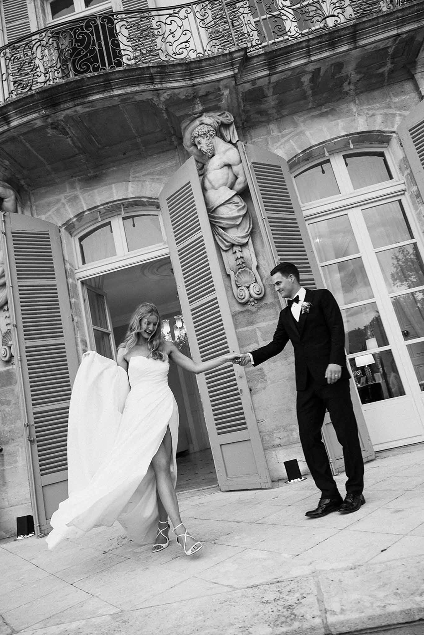 Black and white of couple holding hands stepping through shuttered doors onto chateau terrace with stone relief sculptures