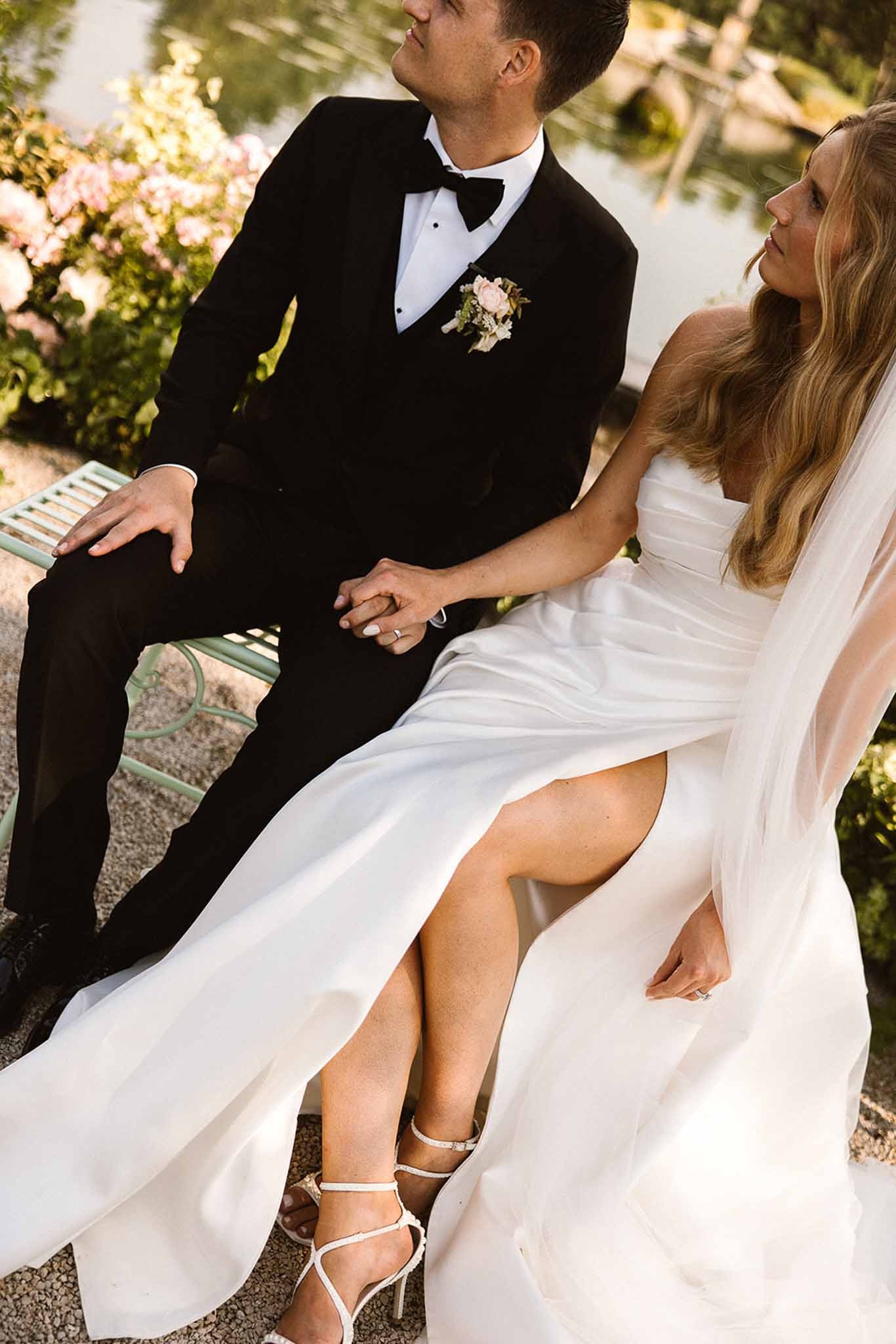 Bride and groom seated on mint green iron bench in garden with pink roses and water backdrop