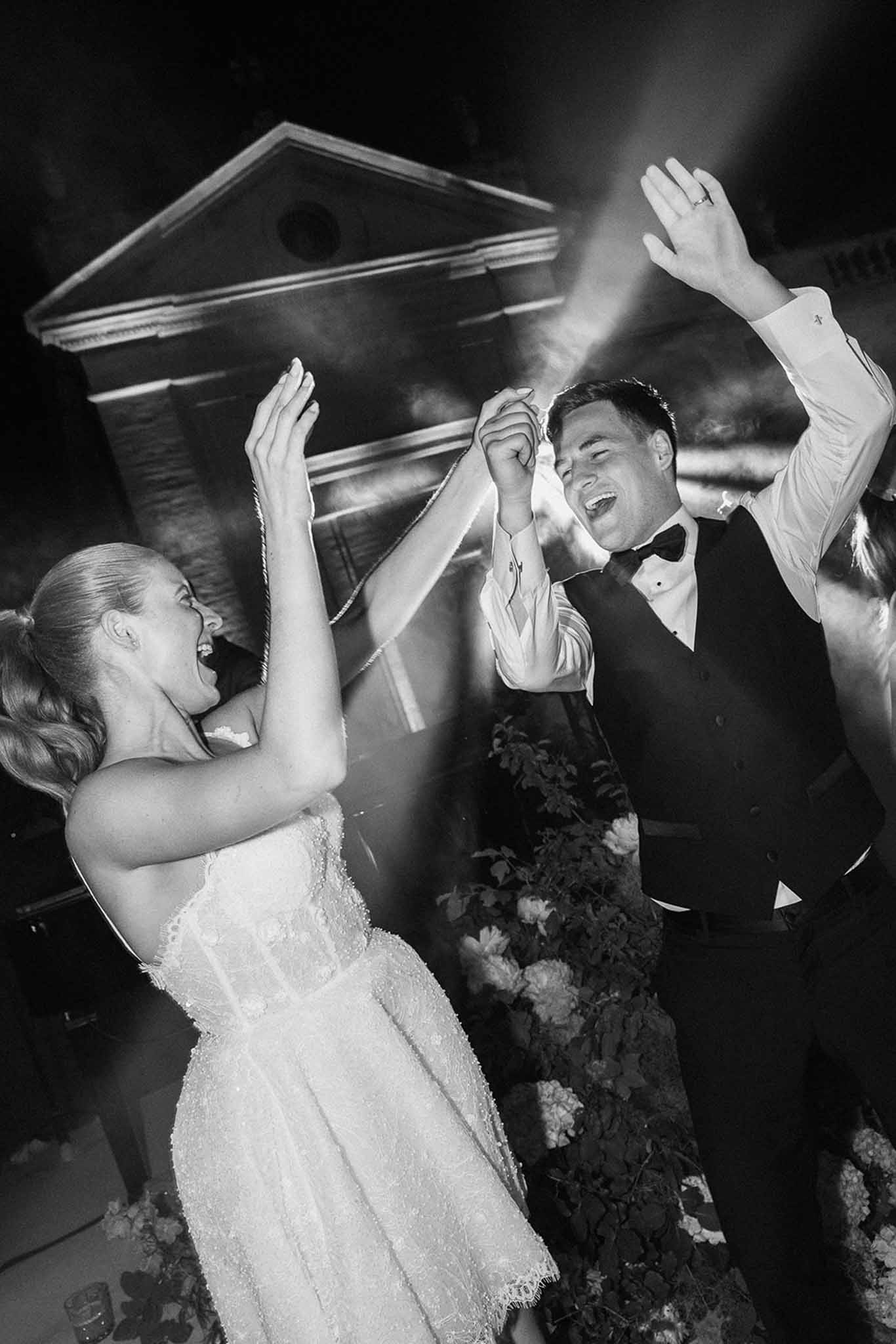 Black and white bride and groom dancing with raised arms under dramatic spotlights at evening reception