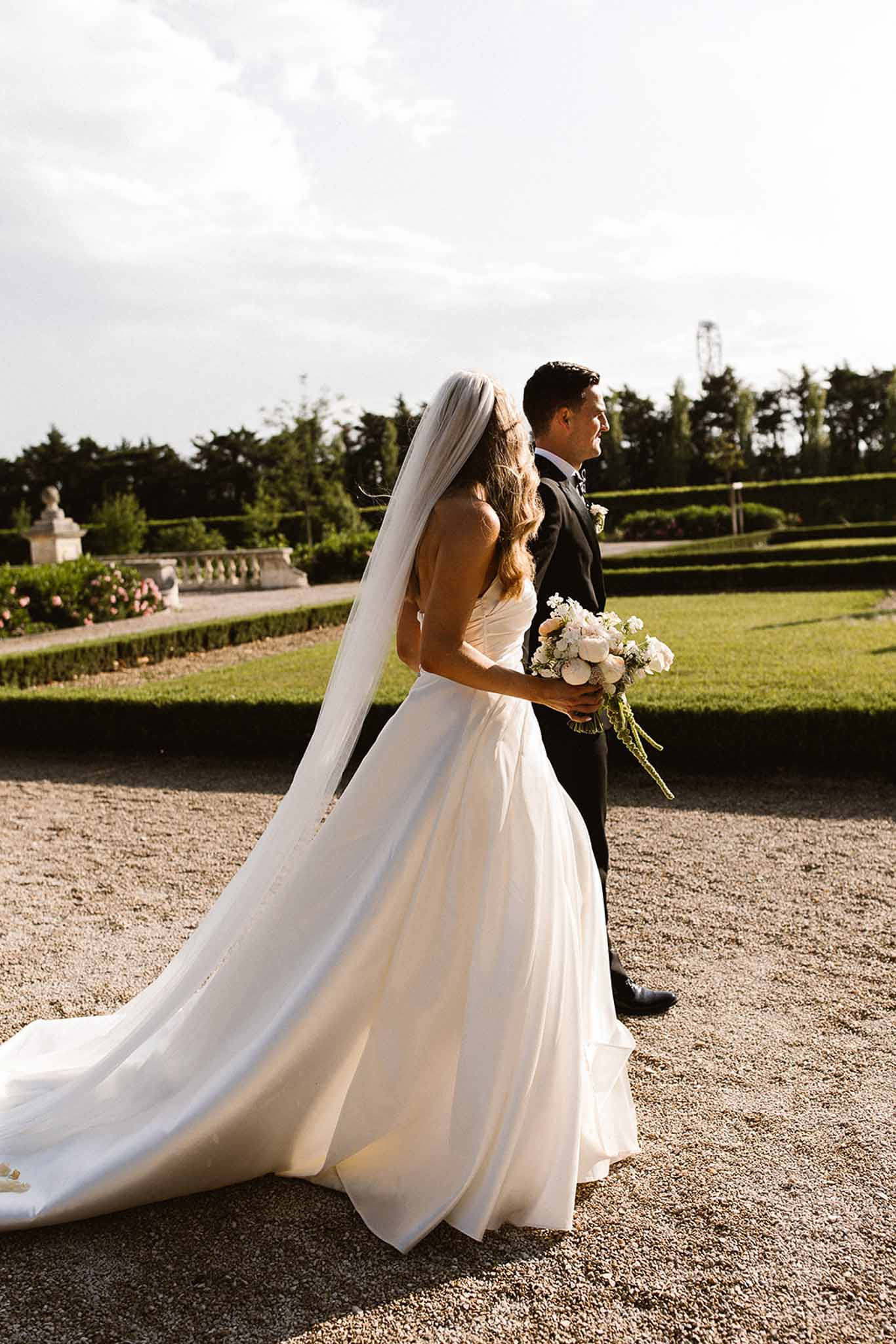 Couple walking on gravel path in formal garden with white peony bouquet and cathedral veil