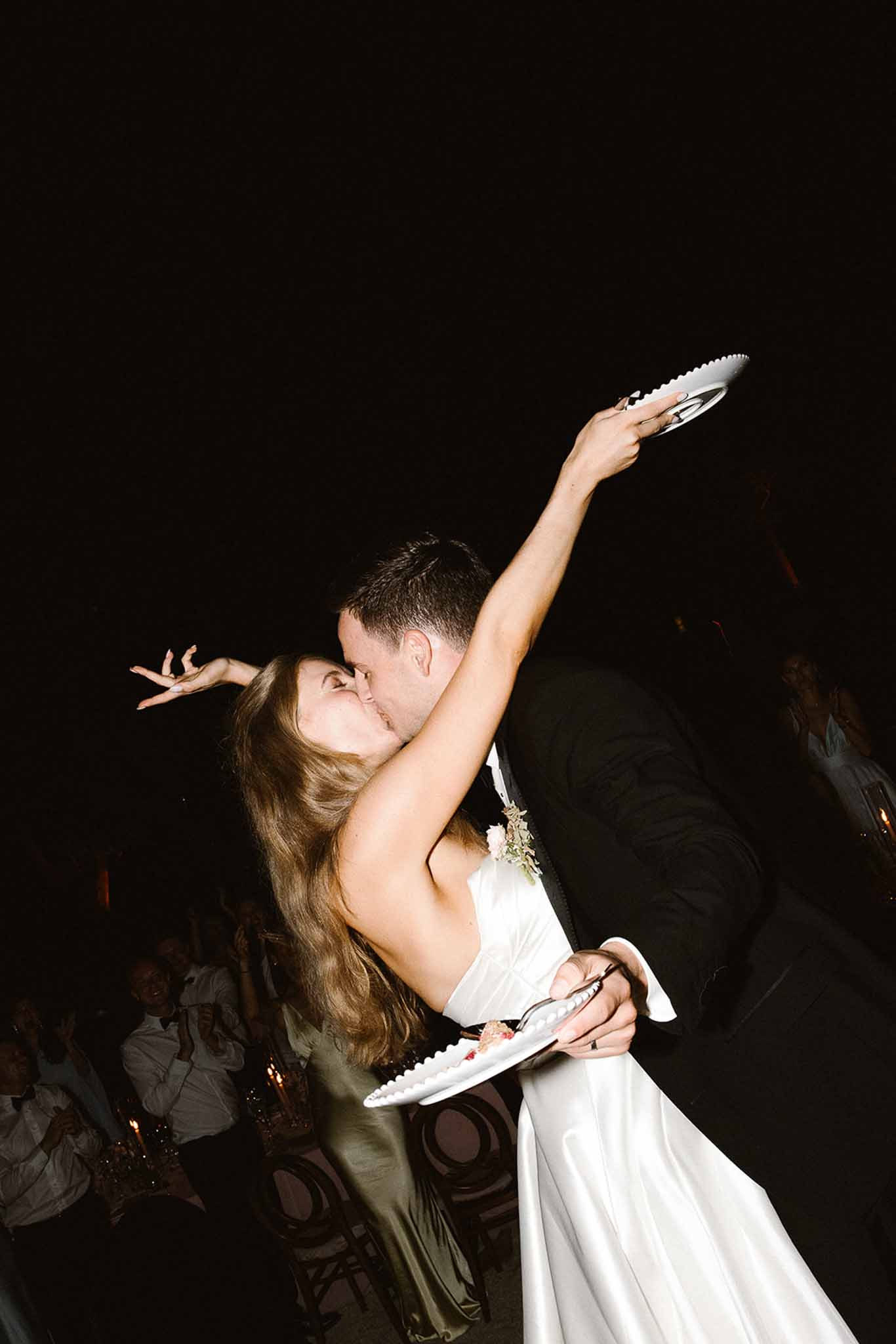 Groom dipping bride mid-kiss holding cake plates aloft during playful evening cake cutting at outdoor reception