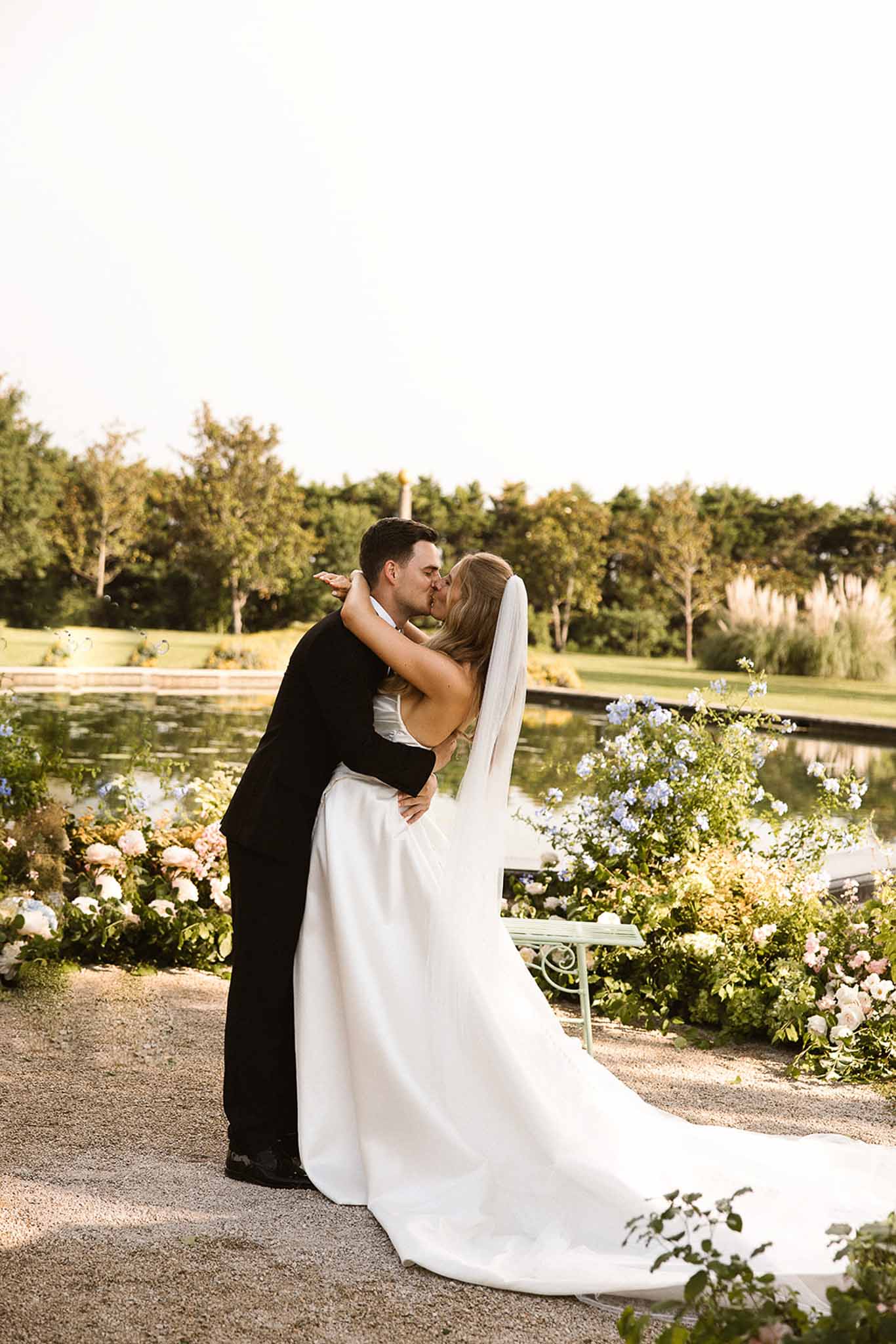 Bride and groom kiss at golden hour beside a reflecting pool flanked by blush rose and white hydrangea arrangements