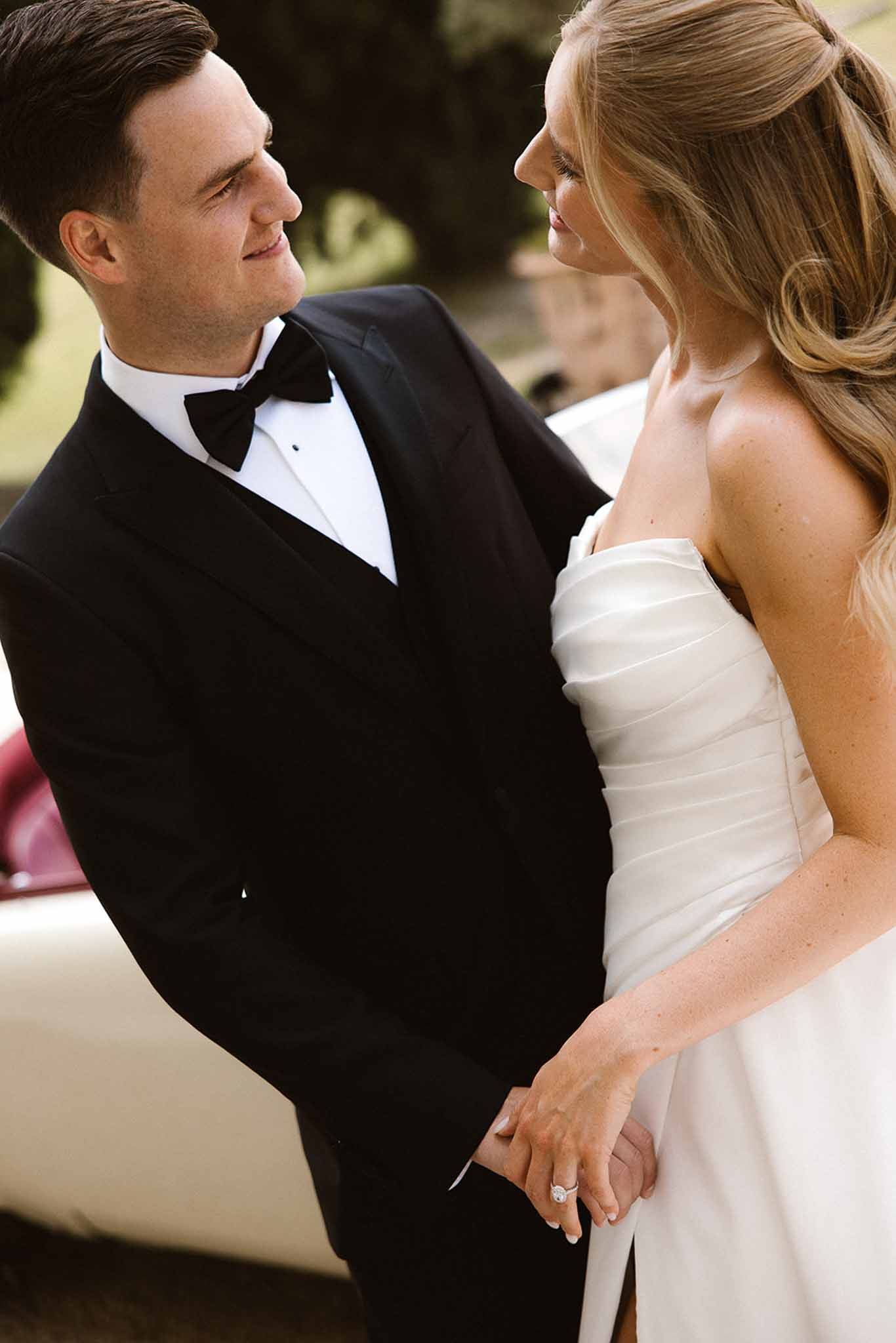 Bride in strapless ivory ruched gown and groom in black tuxedo holding hands with vintage car behind
