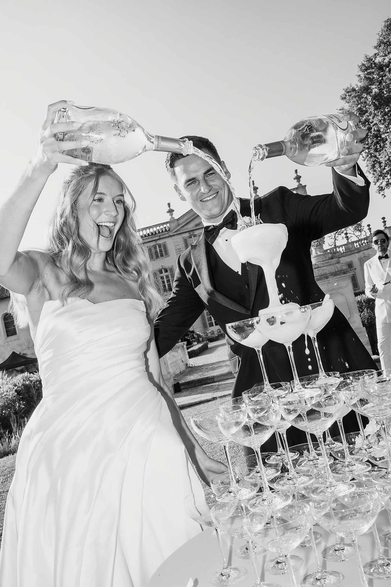 Bride and groom pour champagne into coupe tower before chateau facade in high-contrast B&W