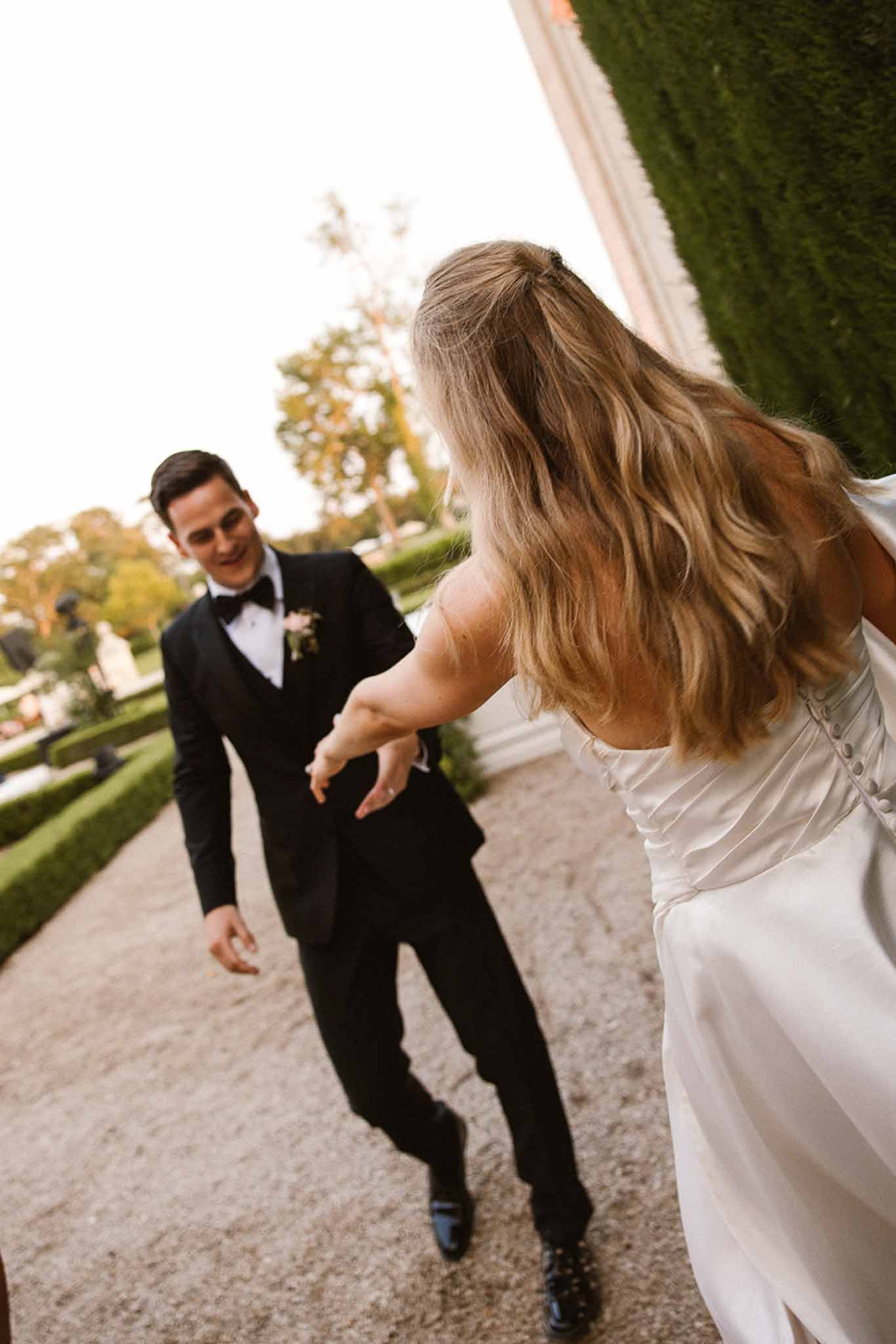 Groom in black tuxedo and bride in white satin strapless gown dancing on gravel path in formal chateau garden