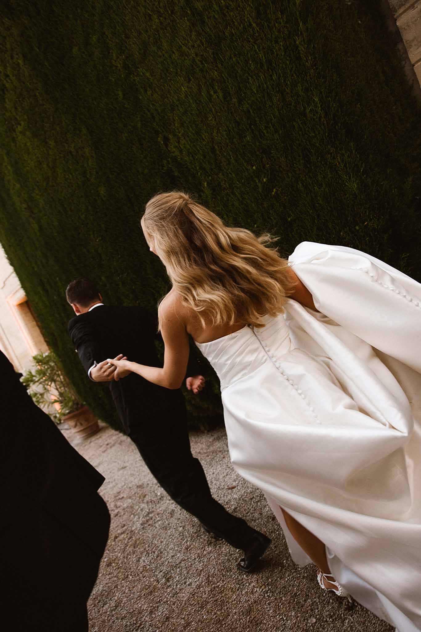 Overhead couple walking along clipped hedge path bride in strapless satin gown with back bow groom in tuxedo