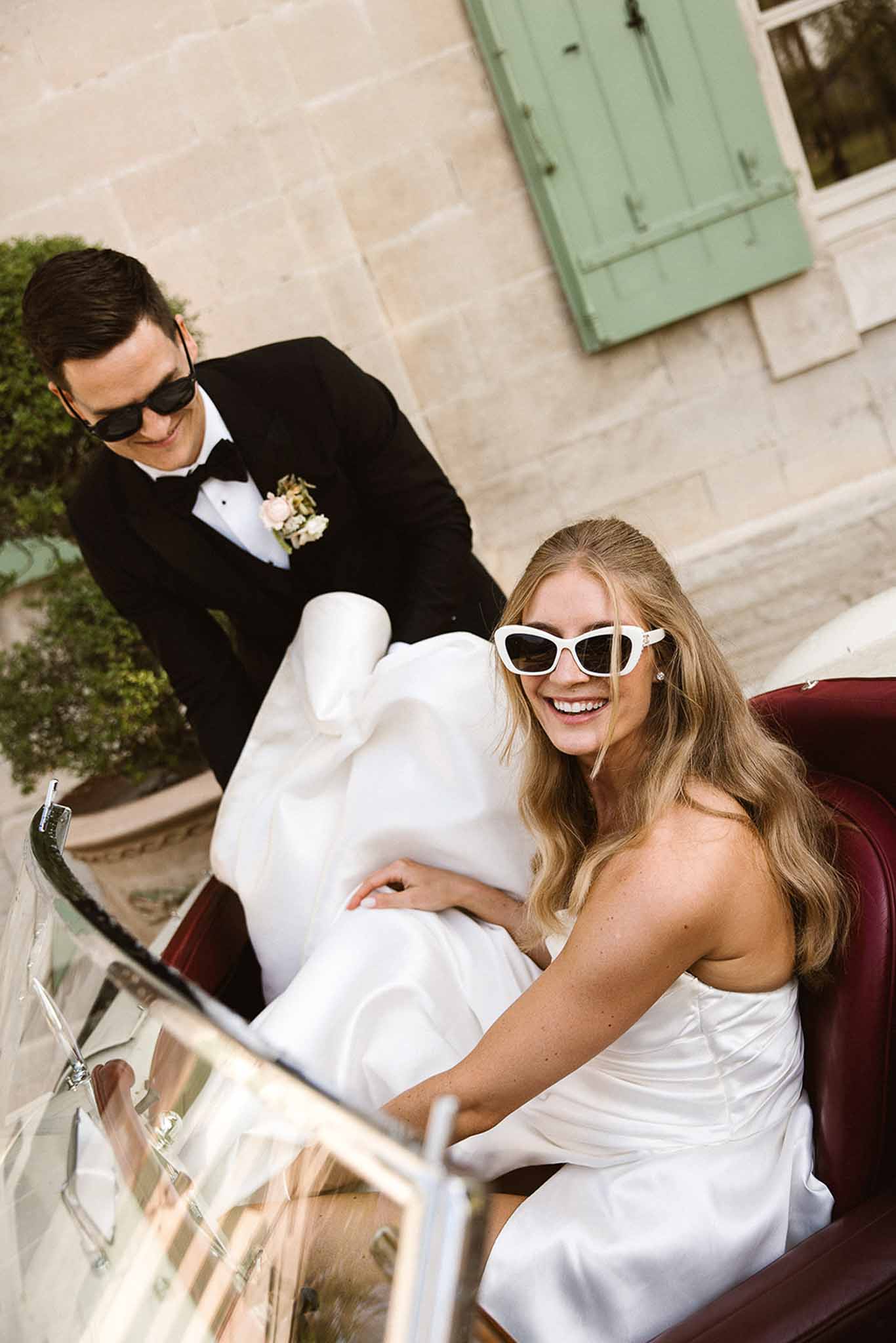 Bride in white strapless gown seated in a vintage car as groom in black tuxedo stands beside her, both wearing sunglasses
