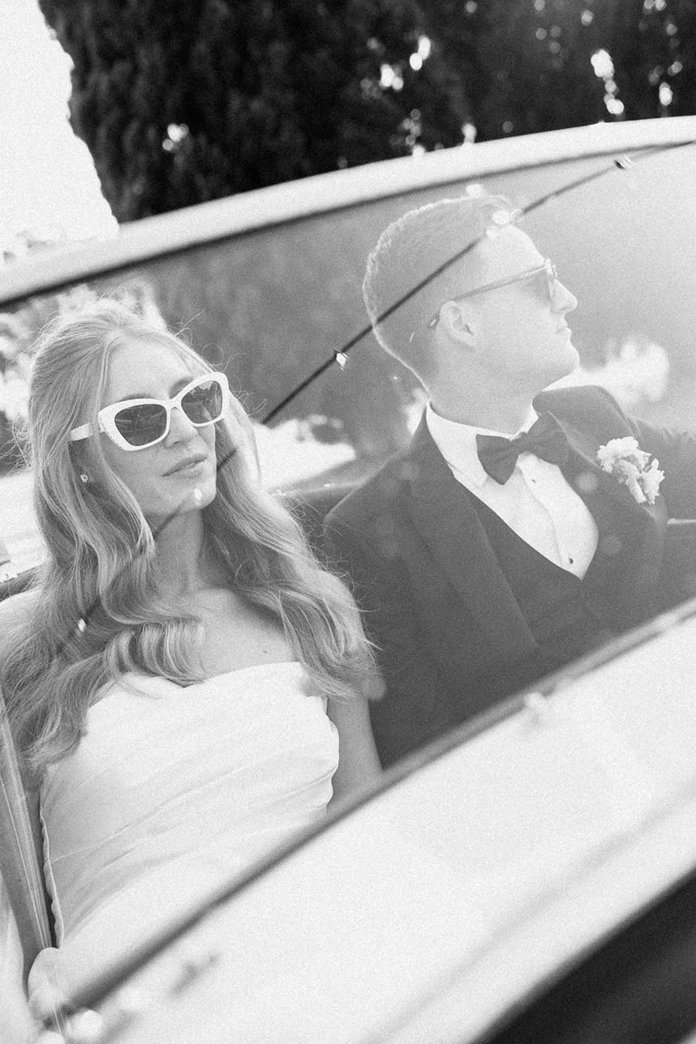Black and white portrait of bride and groom wearing sunglasses photographed through car window