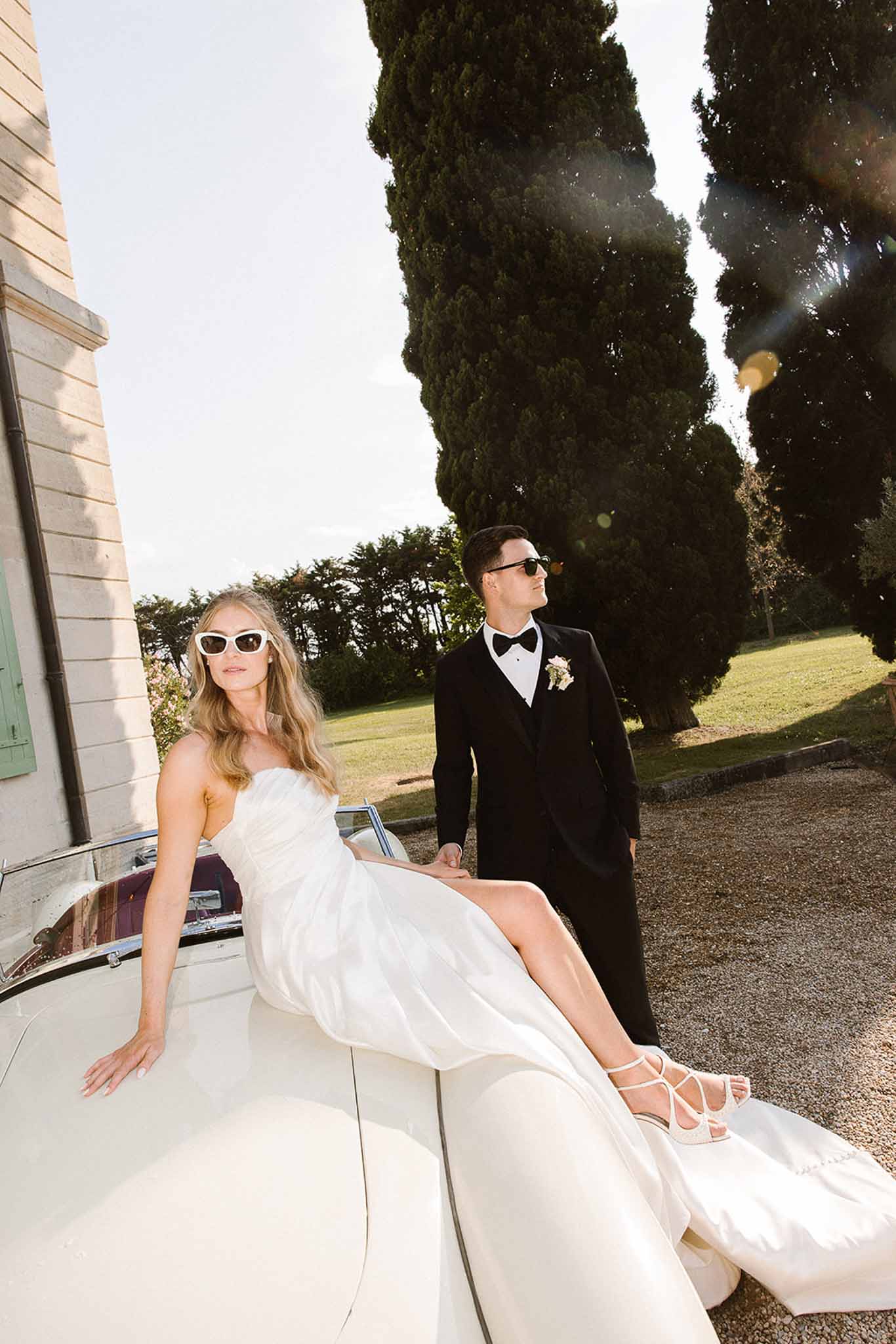 Bride in strapless white gown and groom in black tuxedo posing with vintage white convertible on chateau gravel driveway