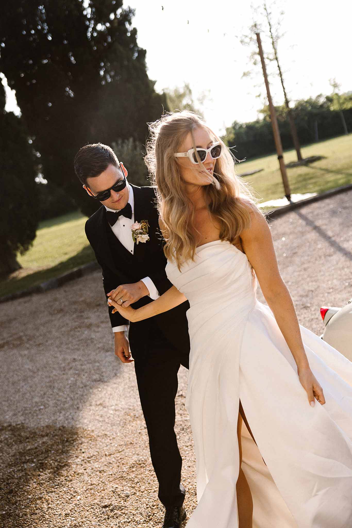 Couple in sunglasses walking arm in arm at golden hour with bride's hair blowing in wind