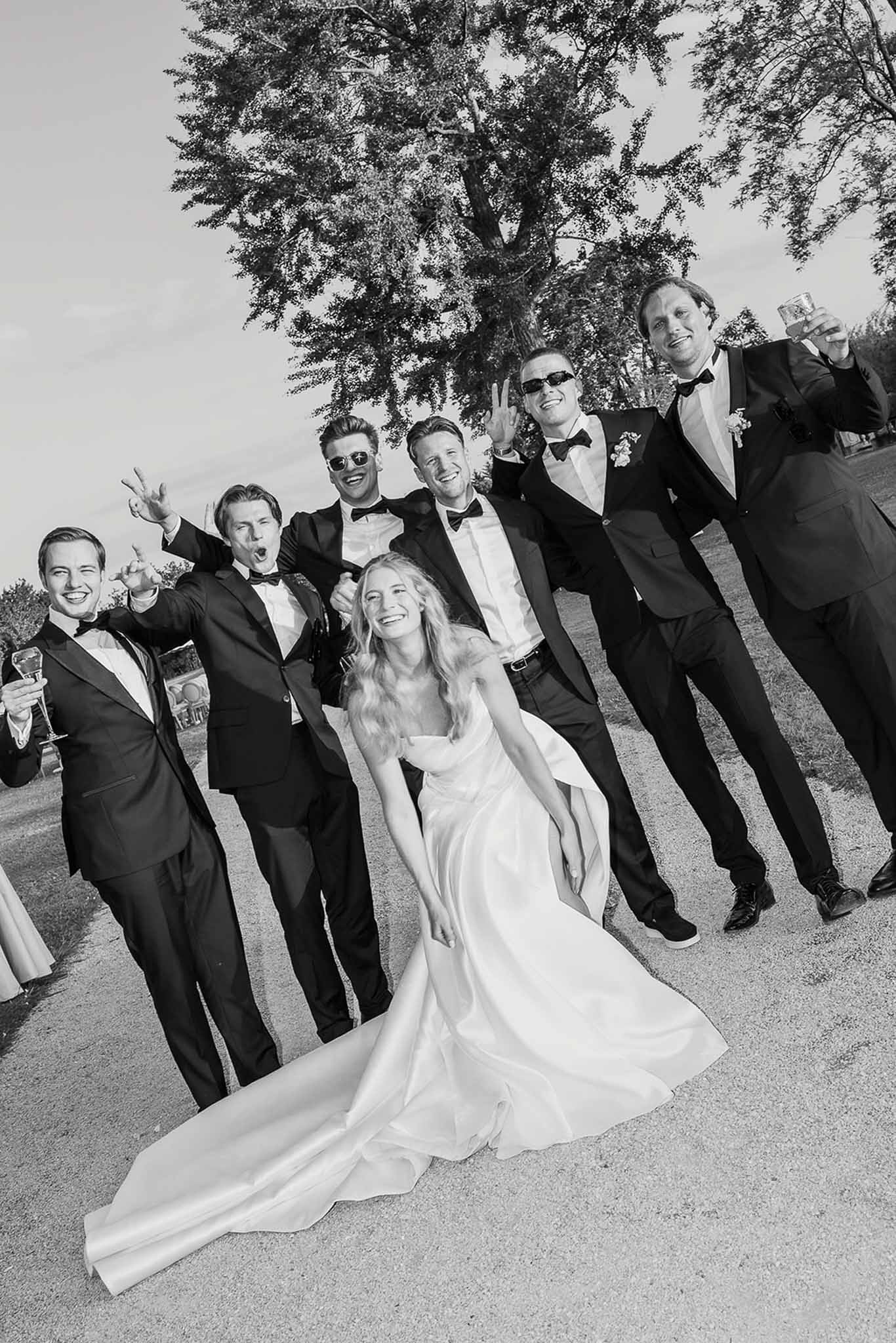 Black and white bride with six groomsmen in tuxedos making playful poses on gravel path