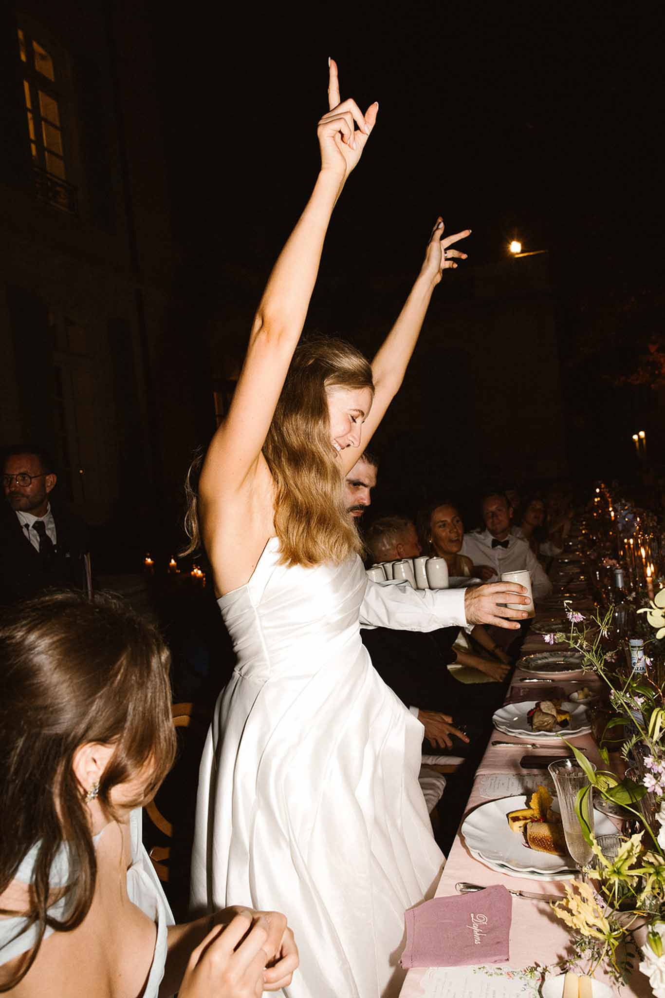 Bride dancing with arms raised beside candlelit table with blush linen and wildflower arrangements