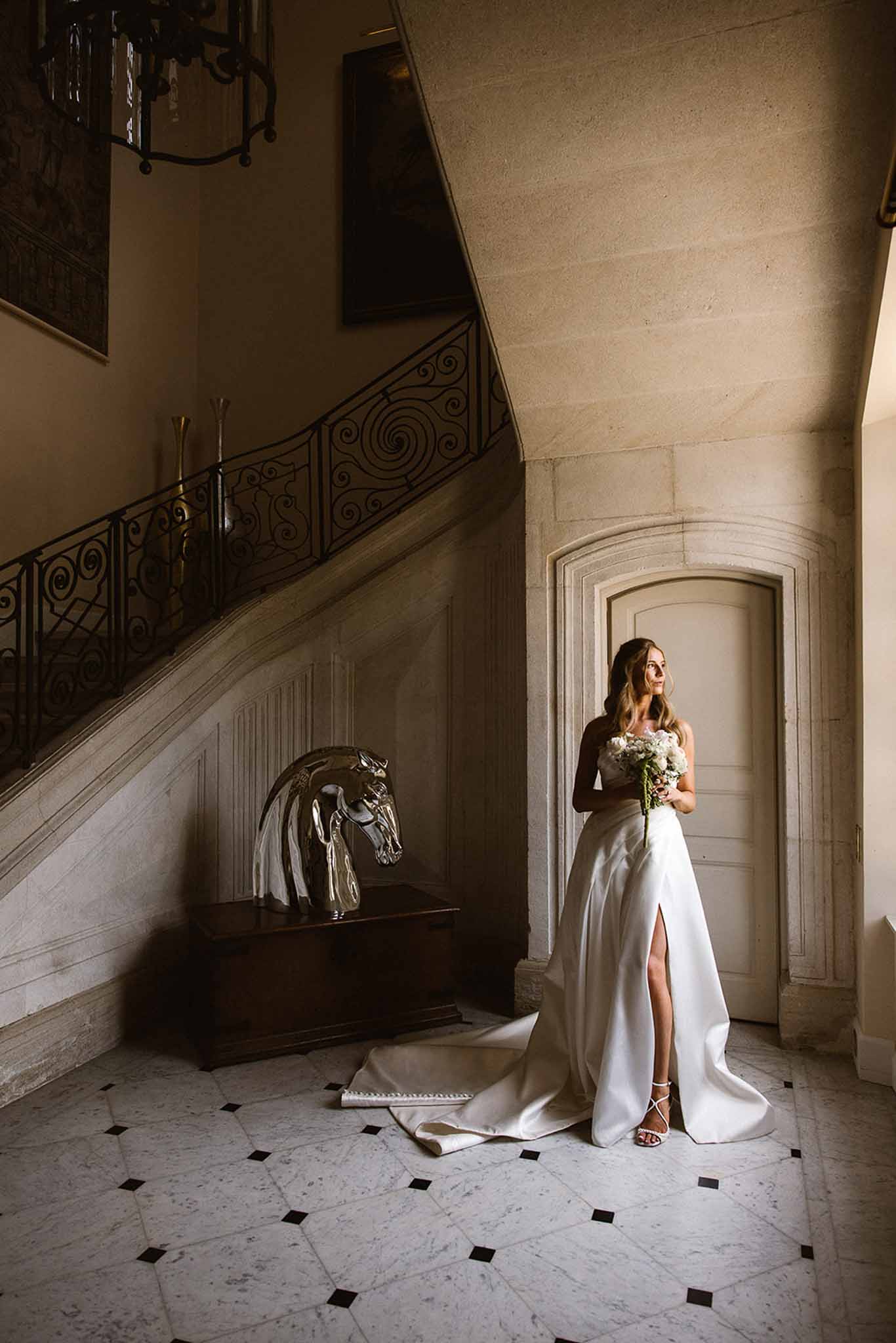 Bride in strapless ivory satin gown holding pink and white bouquet beneath grand stone staircase at French chateau
