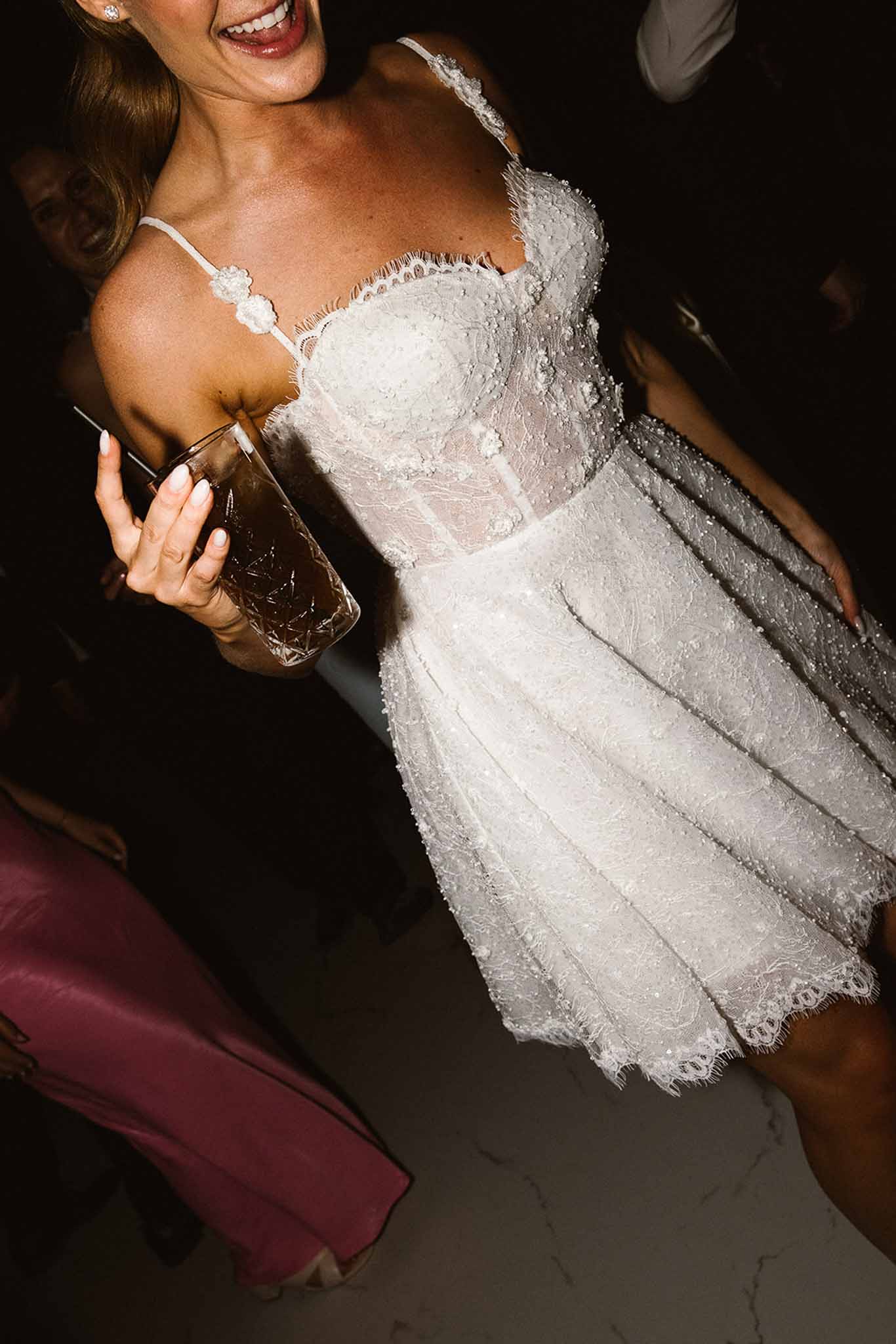 Bride in short white lace mini dress with spaghetti straps laughing and holding cocktail at evening party