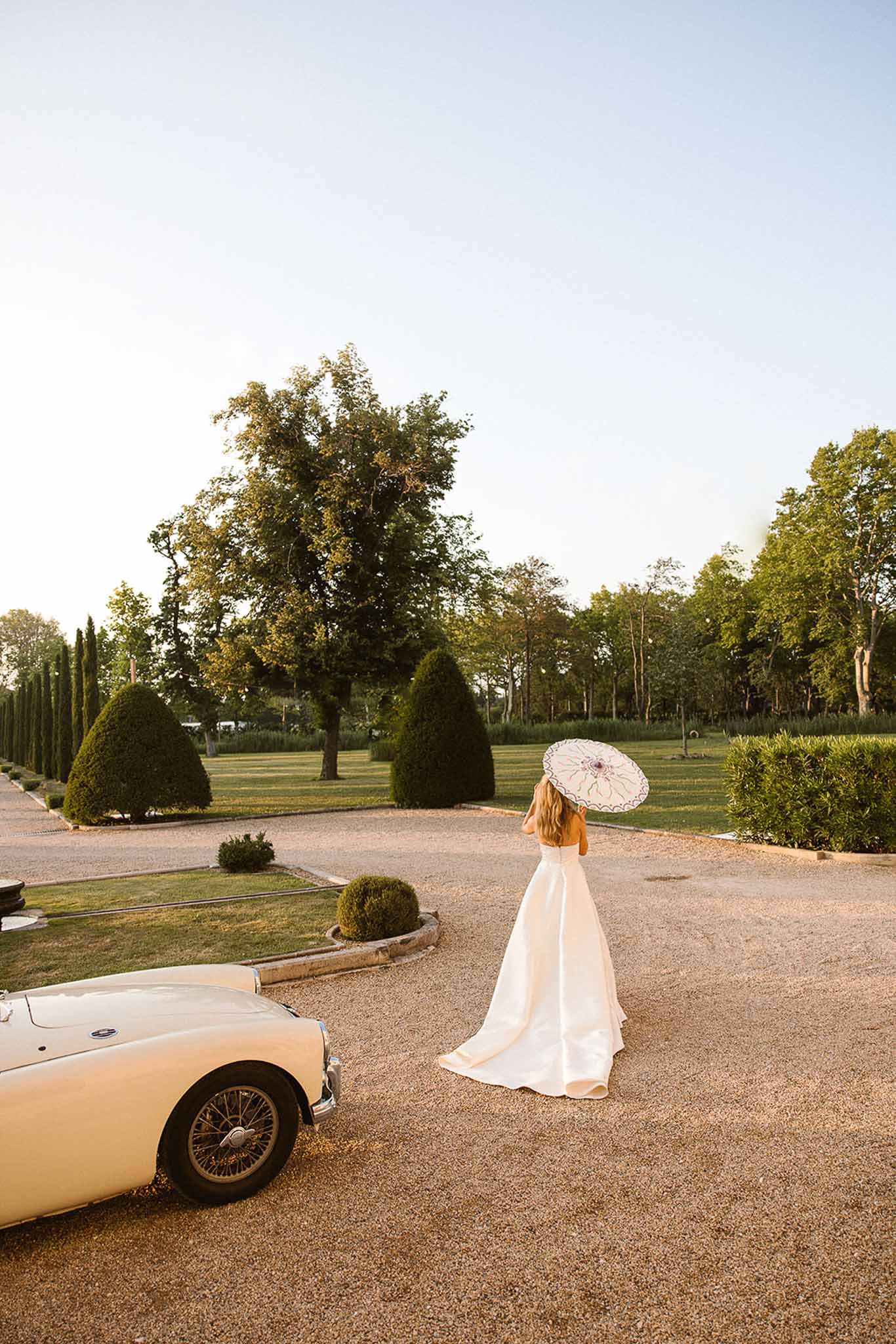 Bride in white satin ballgown with parasol beside vintage convertible on cypress-lined chateau driveway at golden hour