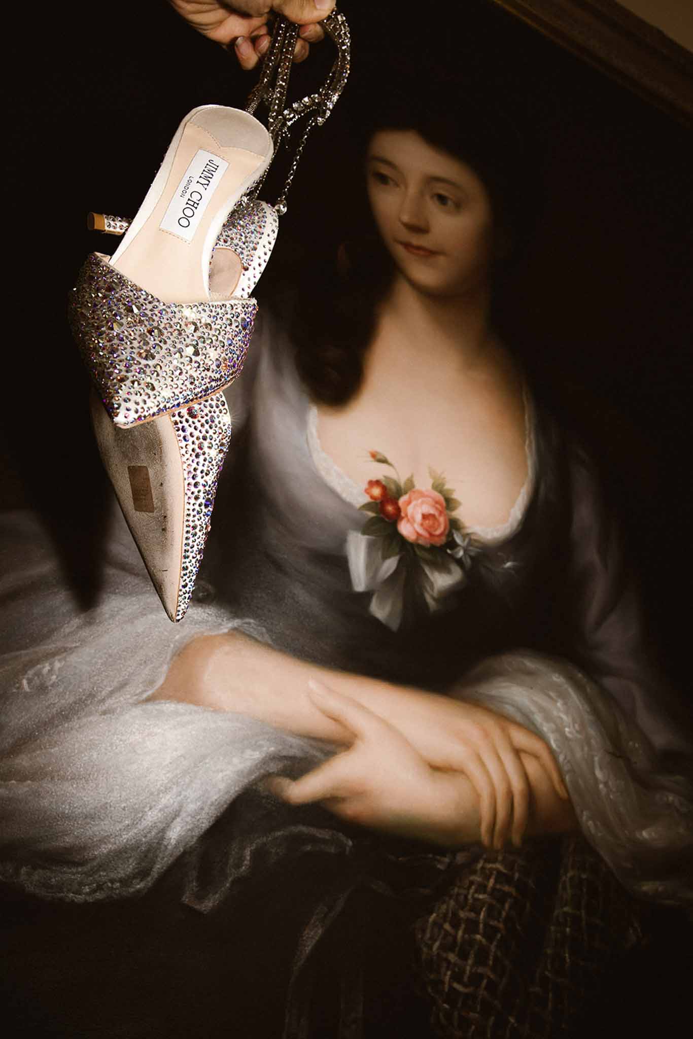 Crystal-embellished Jimmy Choo bridal mules held before antique oil painting portrait in chateau interior