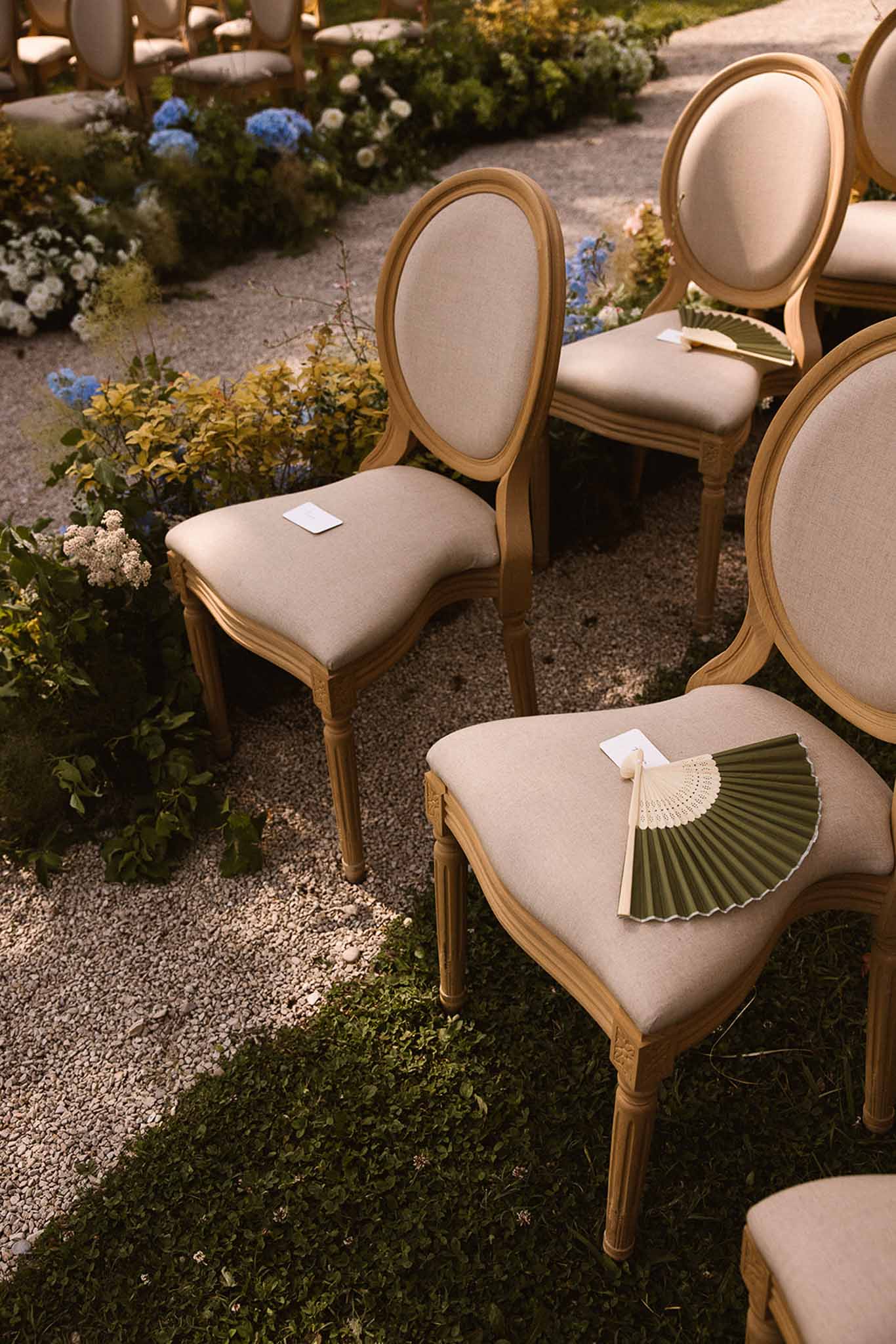 Ceremony chairs with olive green lace fans and white cards on linen seats flanked by blue hydrangea garden borders