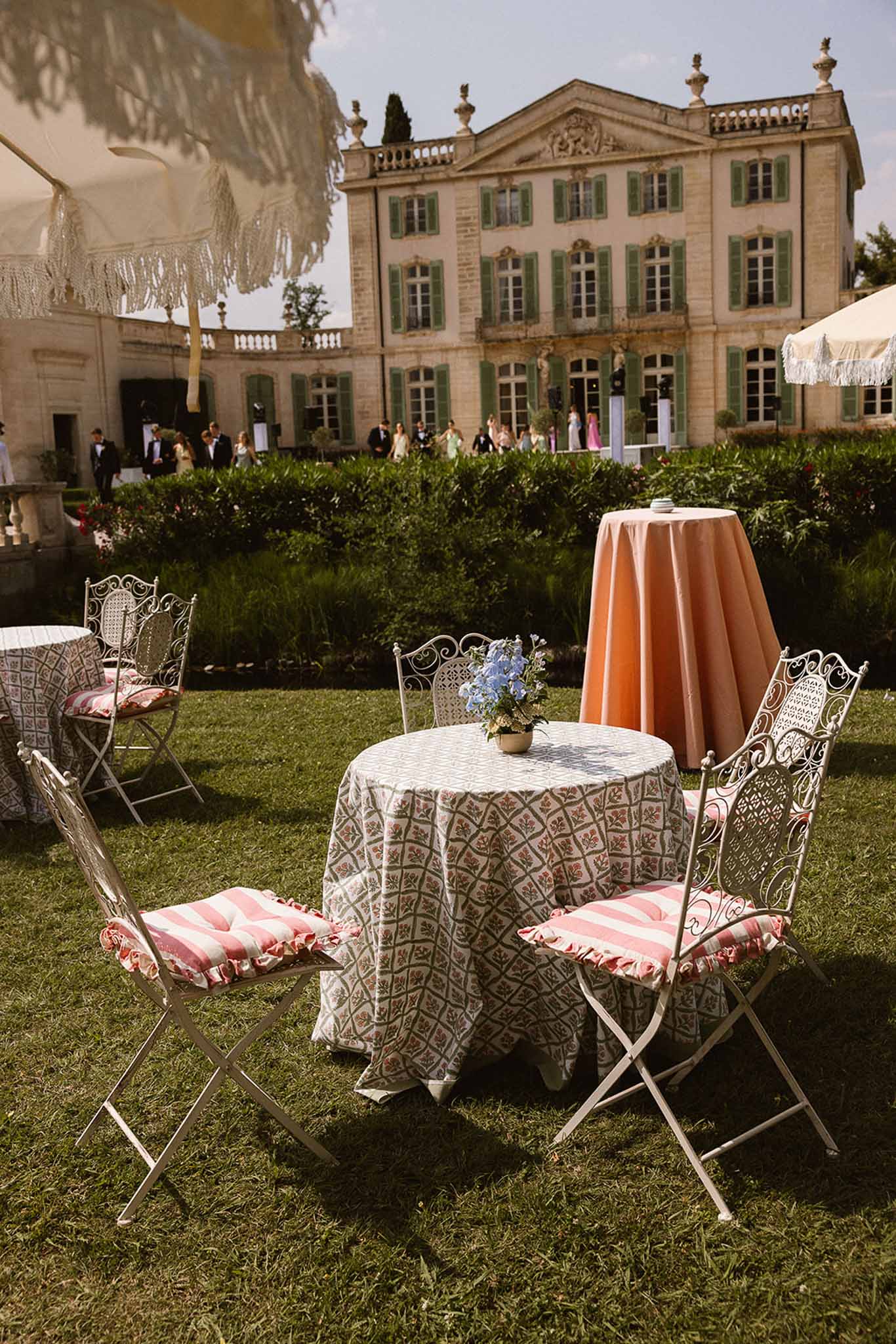 Cocktail hour setup with floral tablecloth and wrought-iron chairs on chateau grounds with guests on terrace