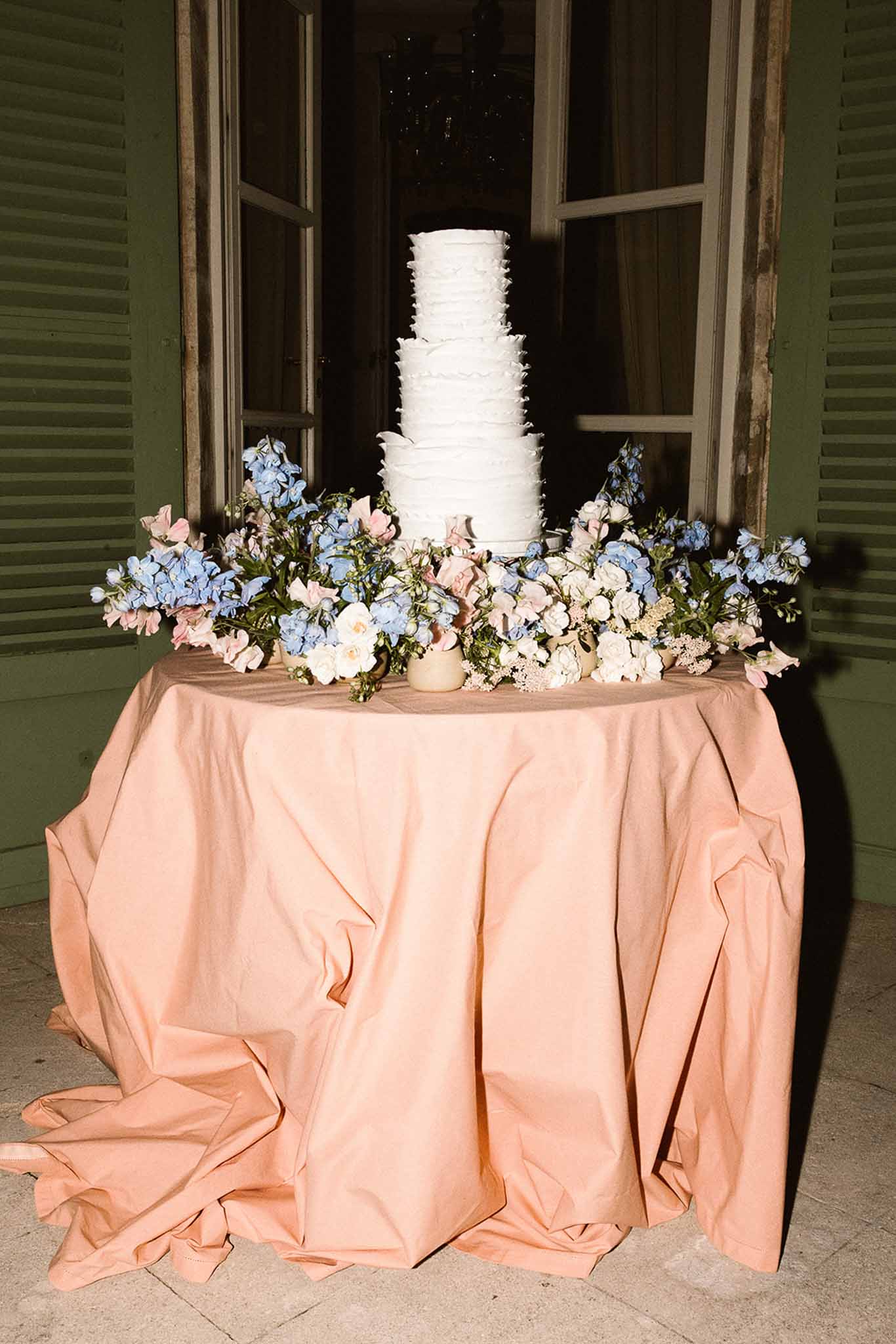 Five-tier ruffle-frosted white cake on peach linen with blue delphinium and blush sweet pea flowers