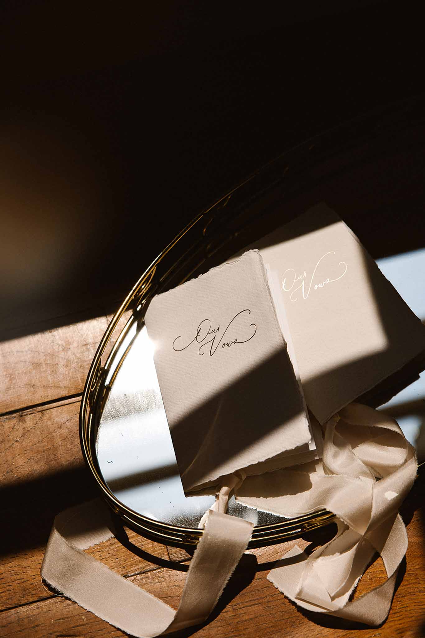 Two deckle-edged vow booklets with calligraphy propped against gold mirror with cream silk ribbon