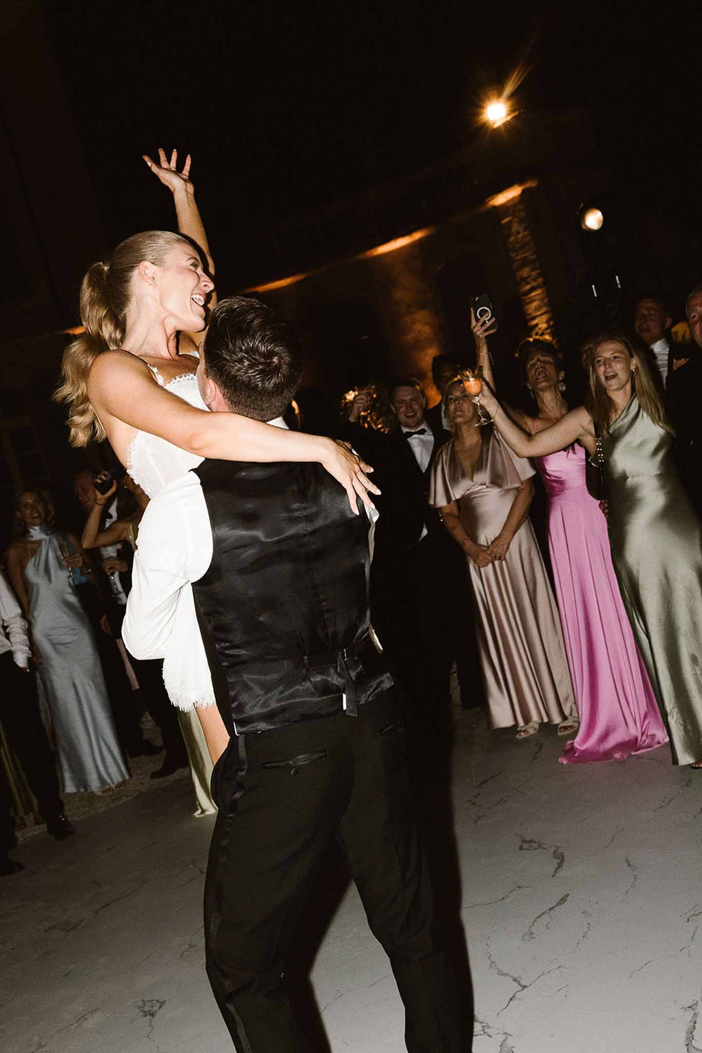 Groom lifts laughing bride in short white lace dress on dance floor as guests film and cheer around them