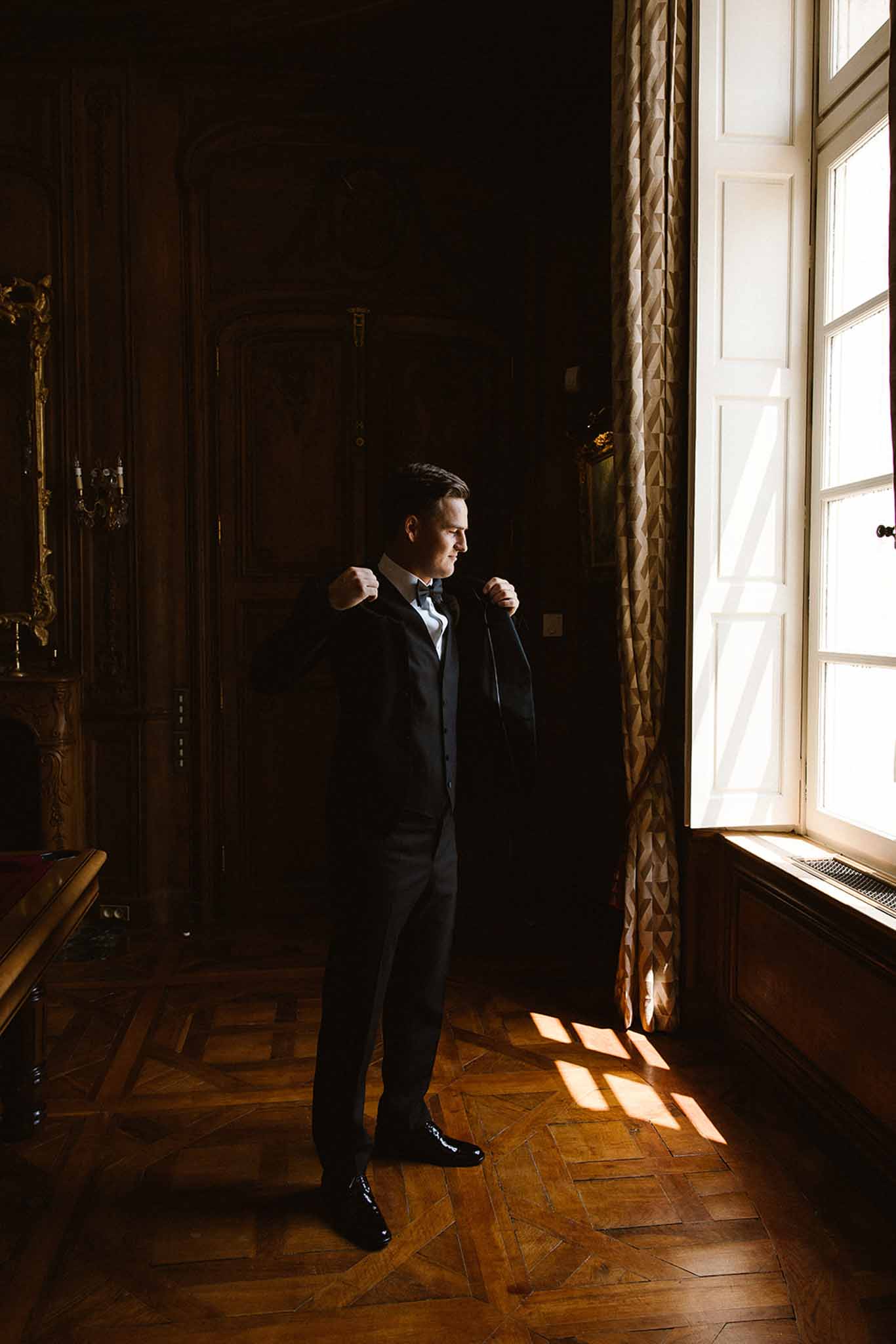 Groom adjusting navy suit lapel beside tall window in wood-panelled chateau room with gilt mirror