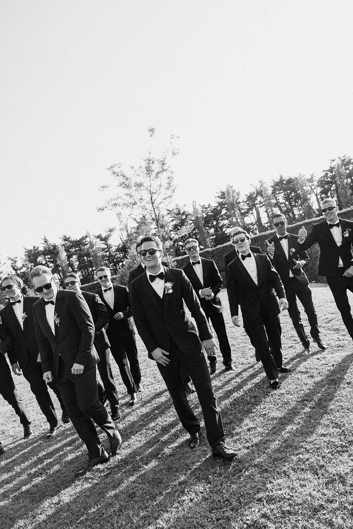 Black-and-white photo of groom and nine groomsmen in tuxedos walking across a lawn wearing sunglasses