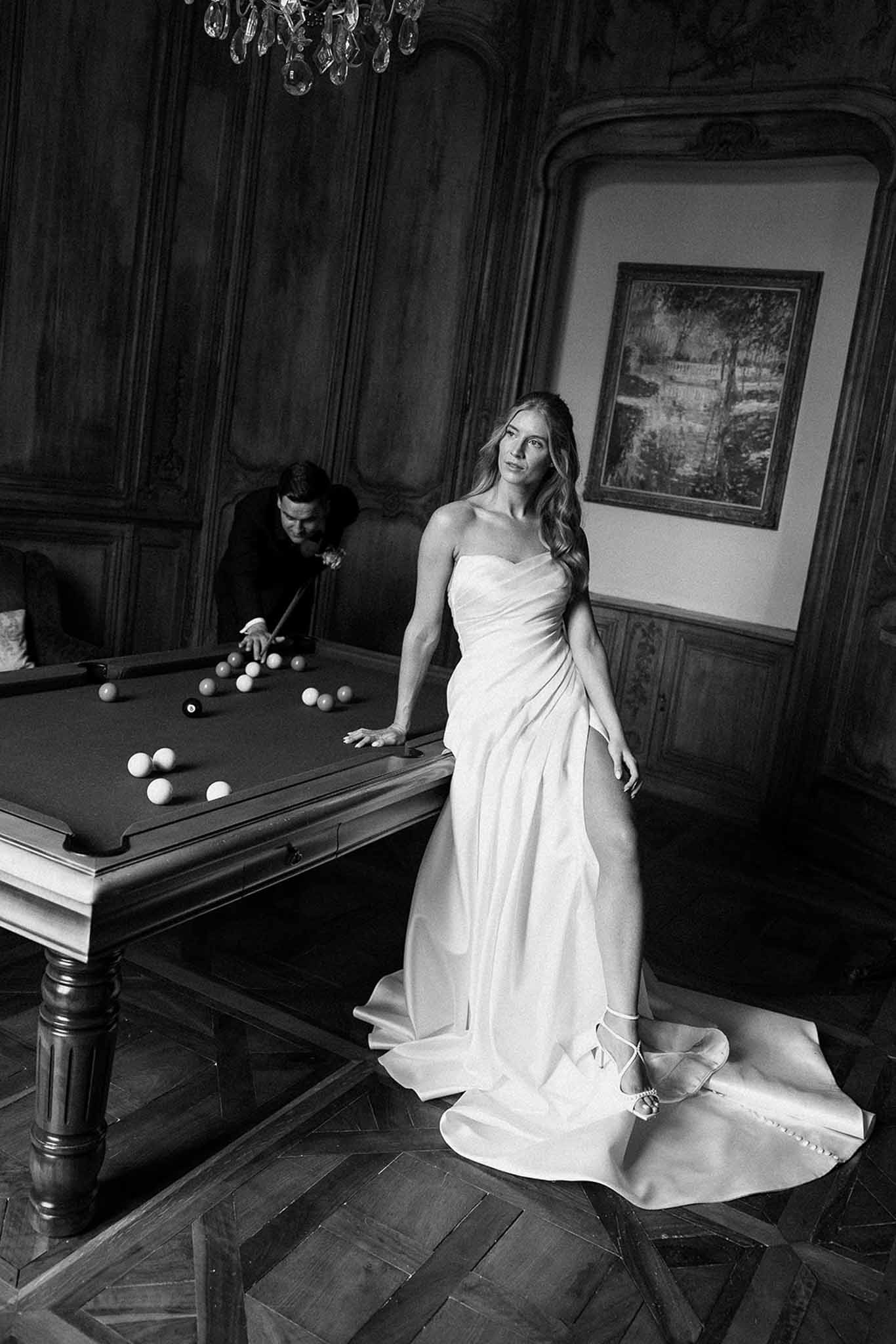 Black and white portrait of bride at billiards table while groom takes a shot in chateau room