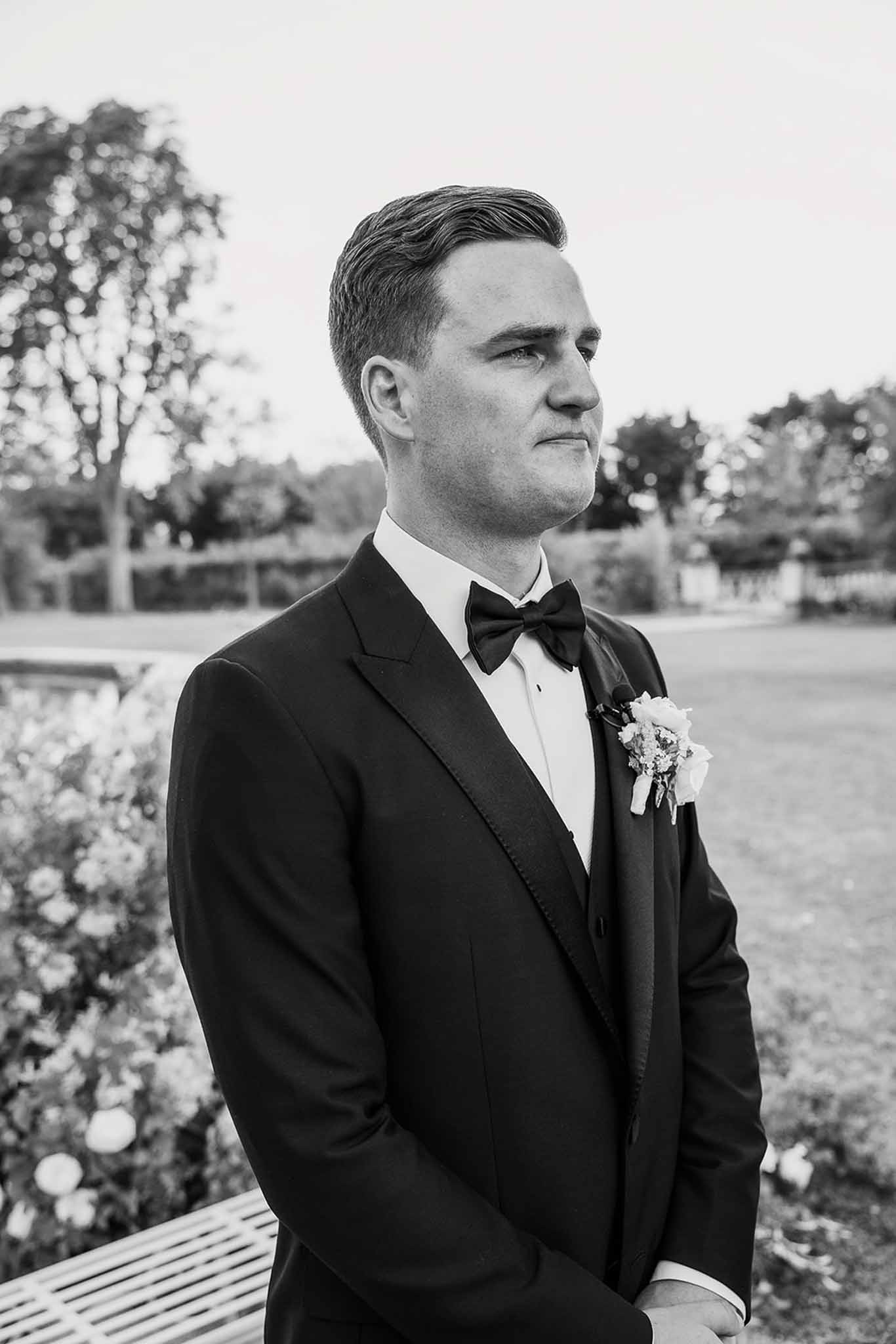 Black-and-white portrait of a groom in a tuxedo and bow tie waiting at the ceremony altar in a formal garden