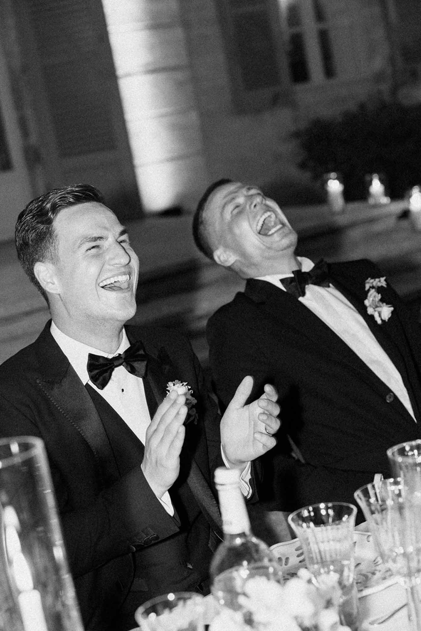 Black and white candid of two men in tuxedos laughing during reception speech at candlelit table