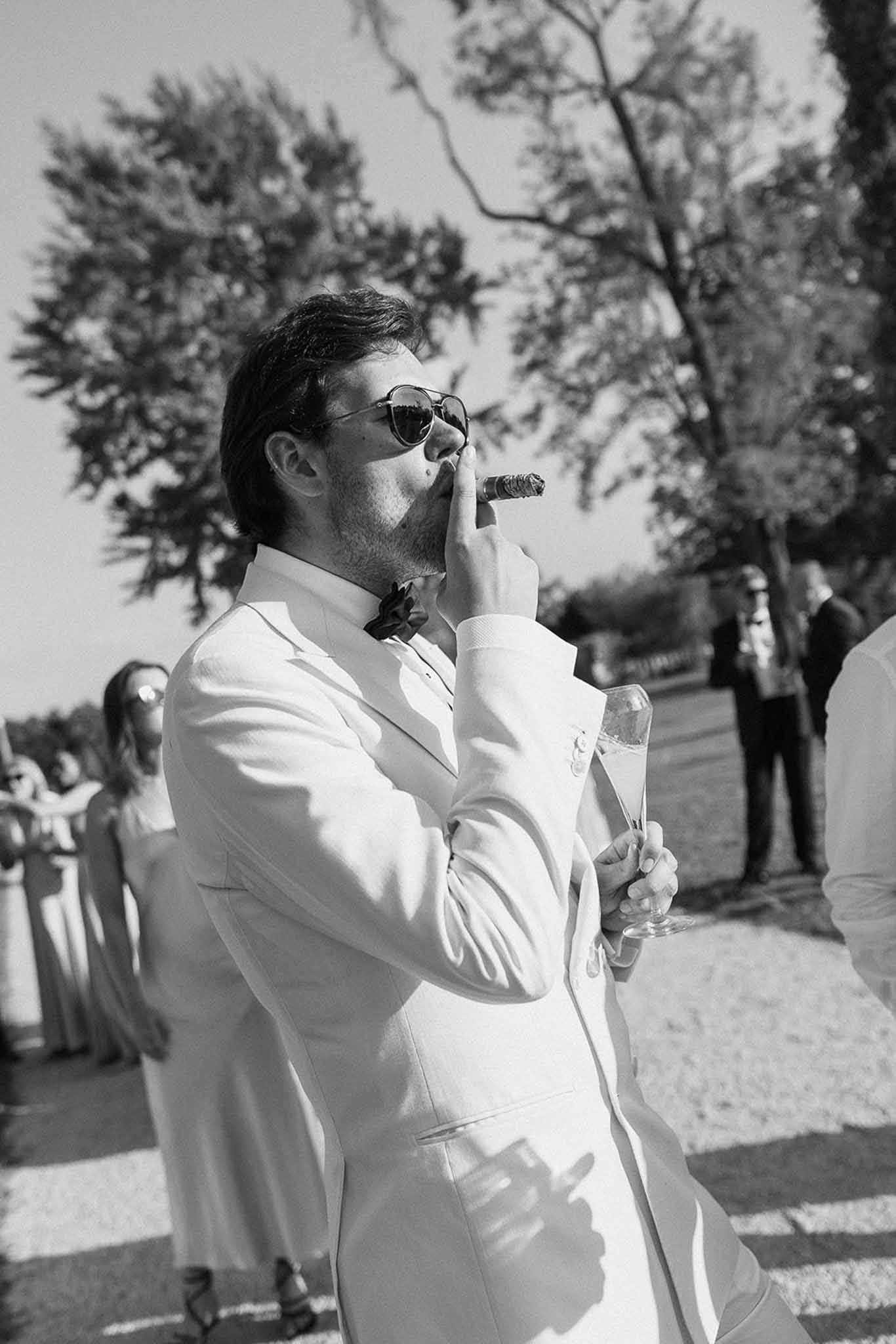 Black and white portrait of groom in white jacket smoking cigar with champagne at cocktail hour