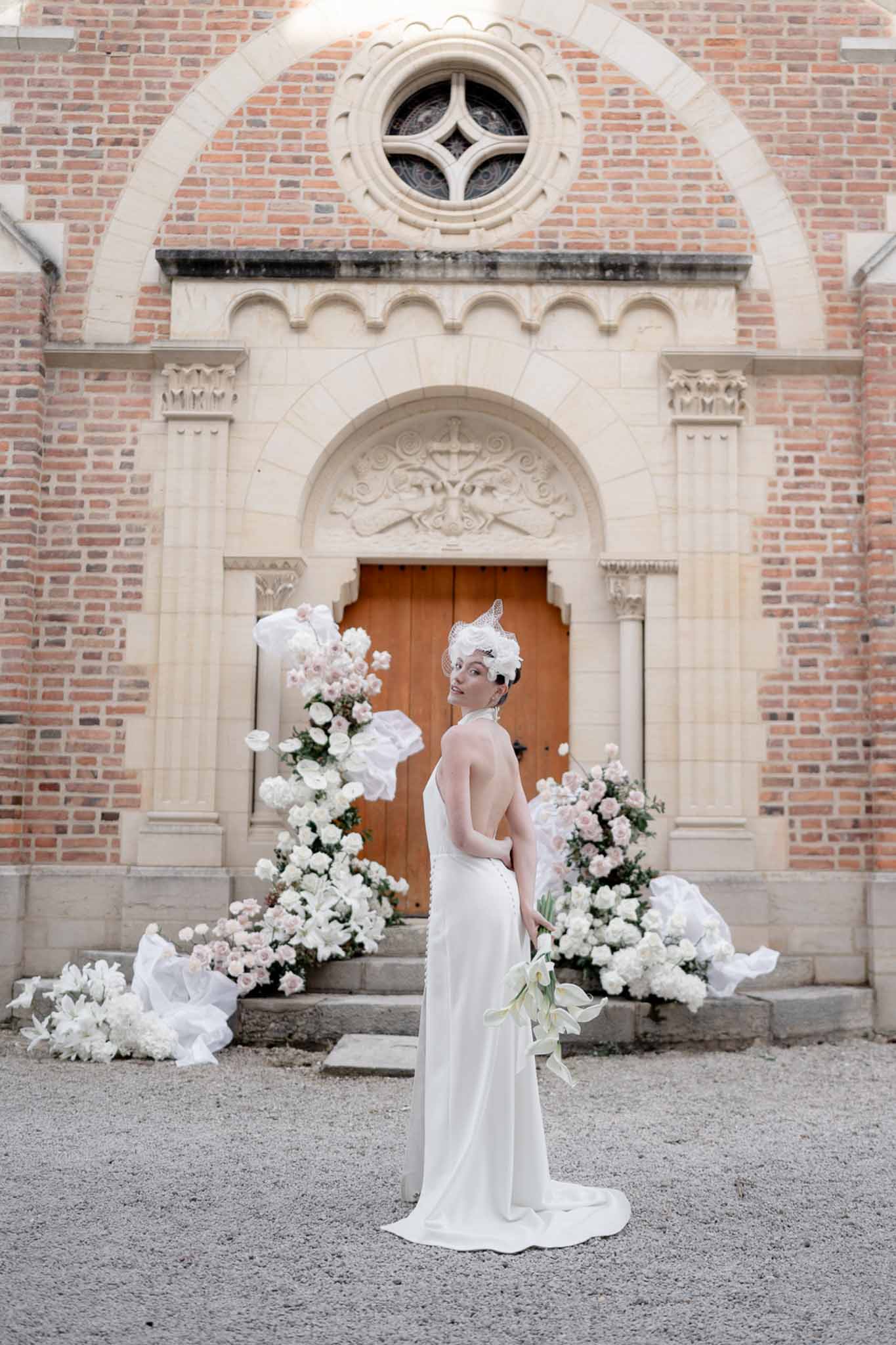 Bride in ivory open-back slip gown with birdcage veil and calla lily bouquet standing before red brick chapel entrance