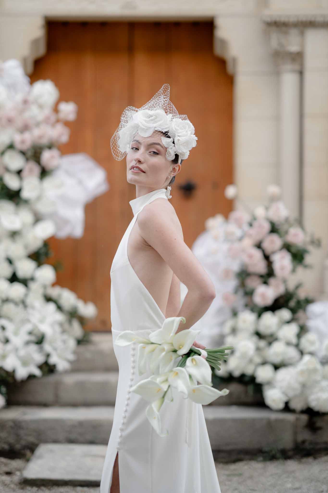 Bride in white halter-neck open-back gown with birdcage fascinator and calla lily bouquet beside rose installations