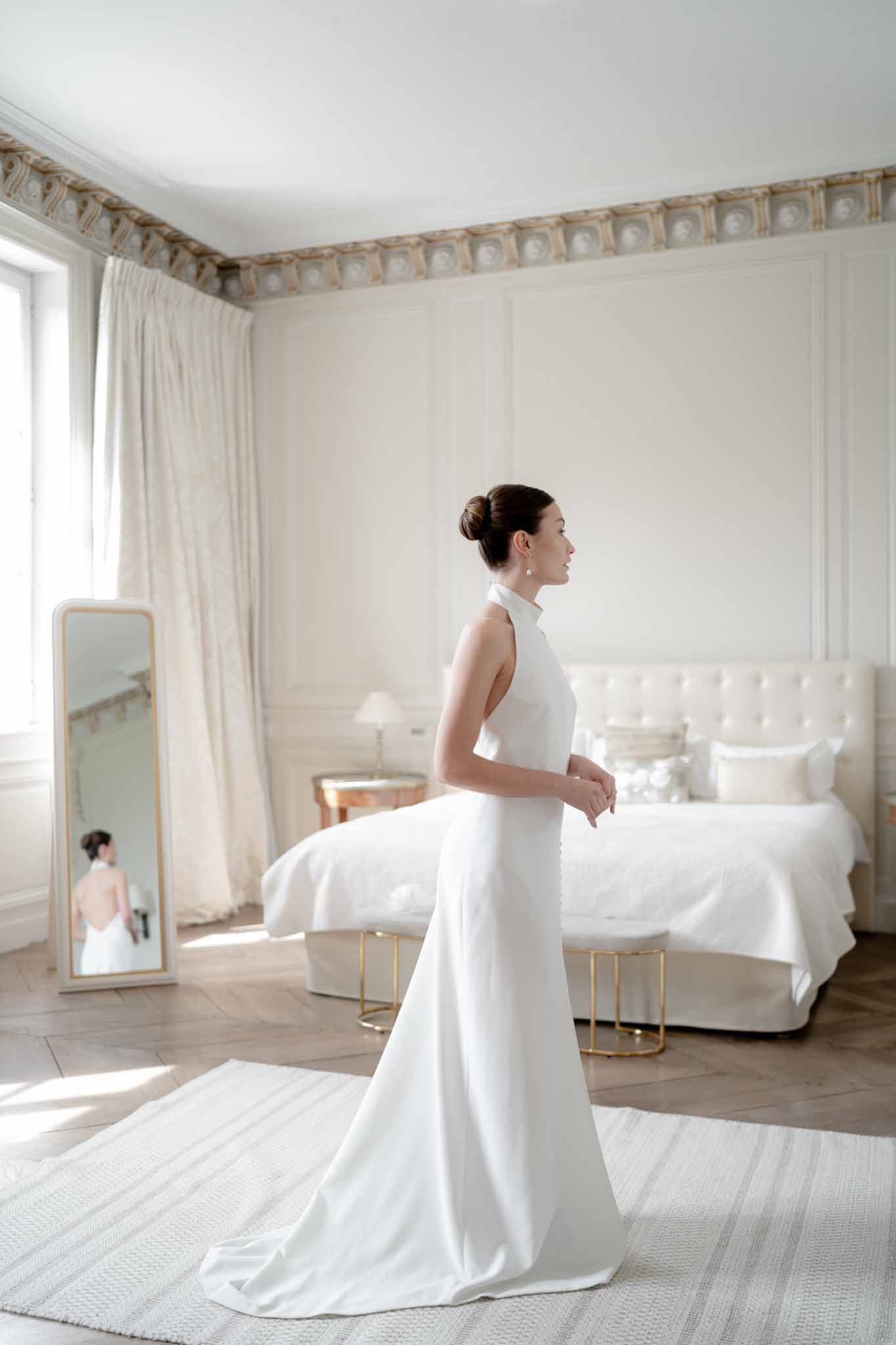 Bride in a white halter gown standing in a panelled chateau bedroom with herringbone floors and a gold-framed mirror