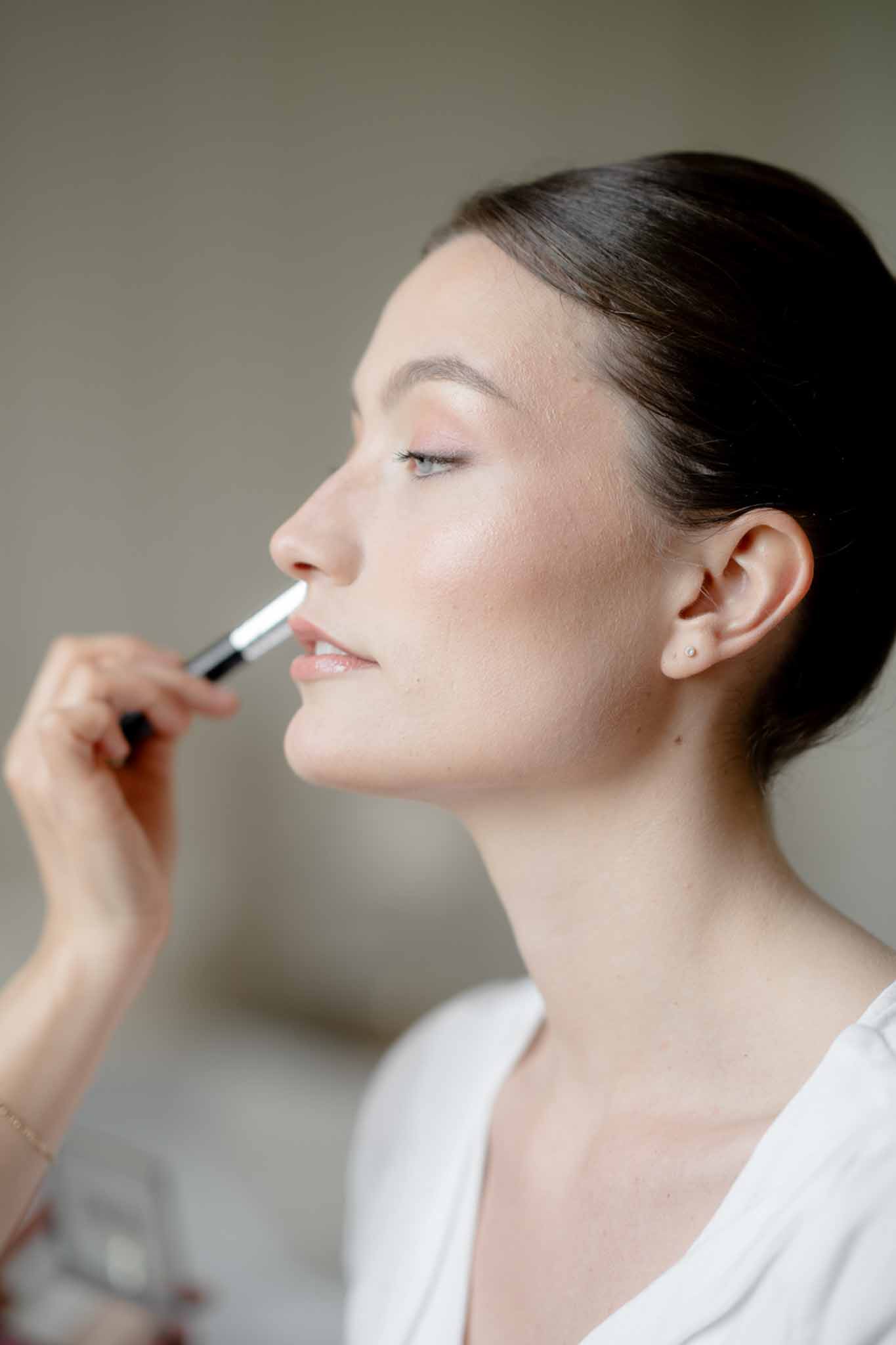 Close-up side profile of bride in white robe having makeup applied by artist with brush to upper lip