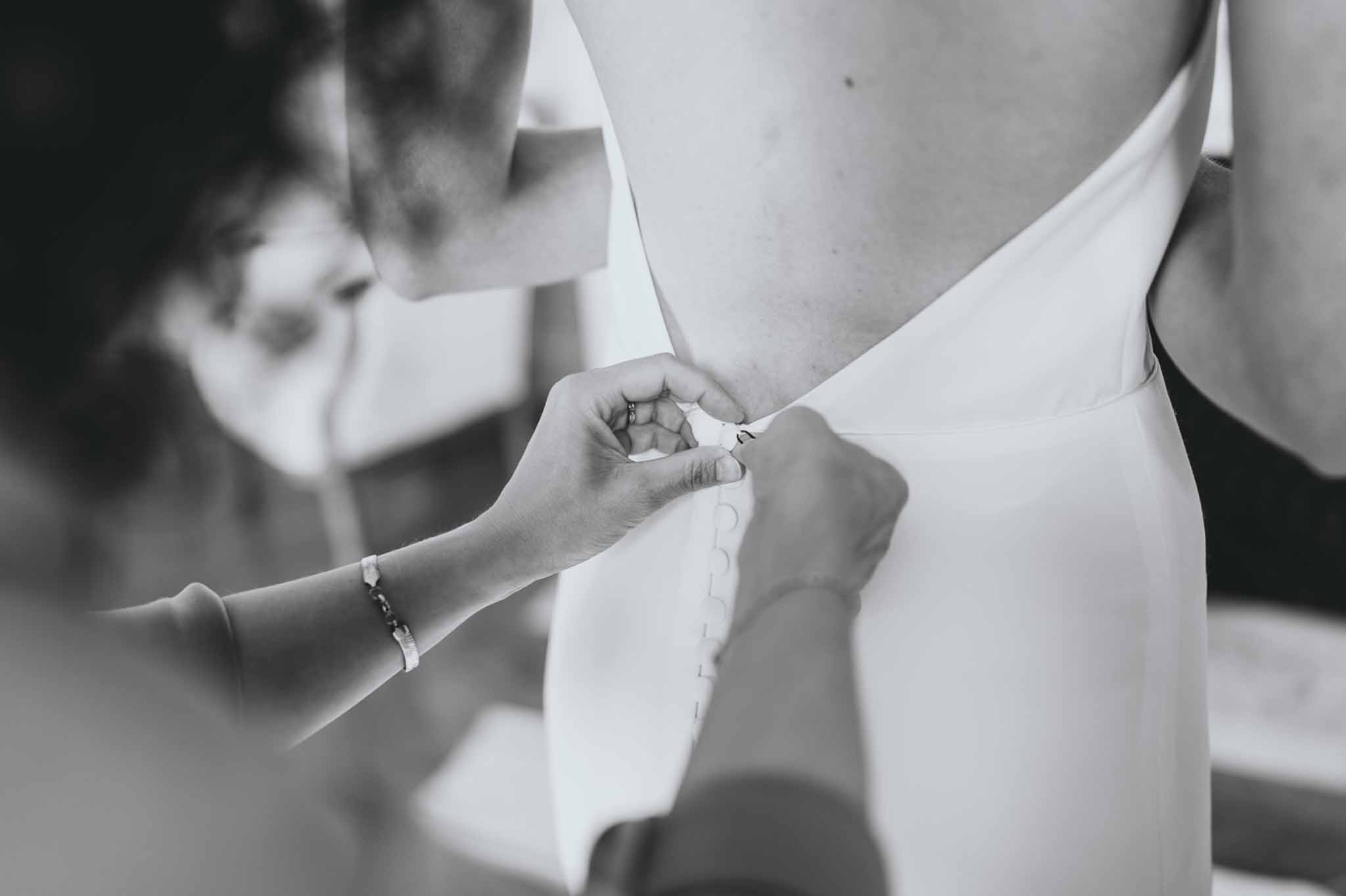 Black-and-white close-up of hands fastening buttons on a bride's fitted wedding dress during getting ready