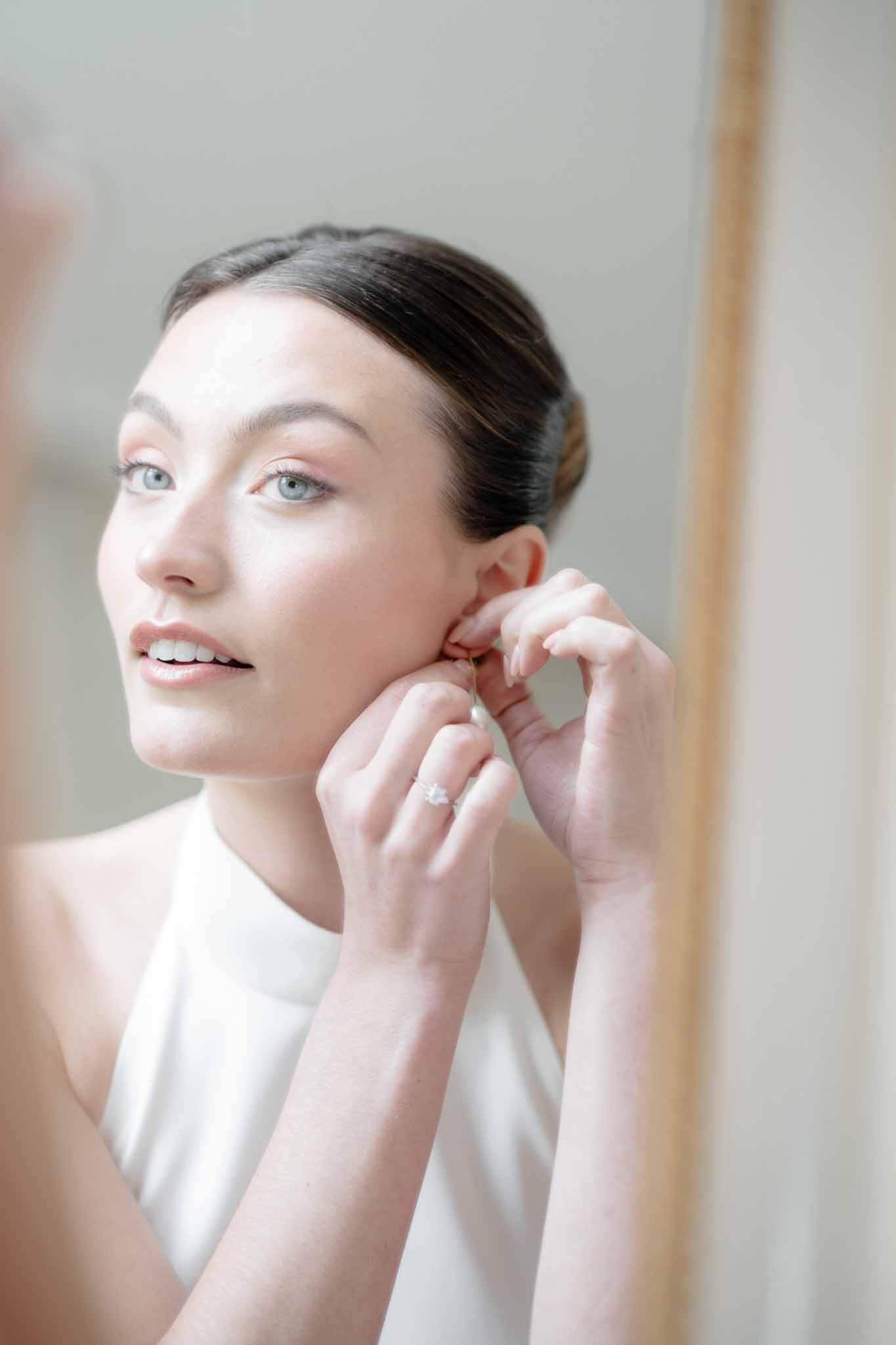 Bride putting on pearl drop earring in wood-framed mirror with sleek updo and diamond engagement ring visible