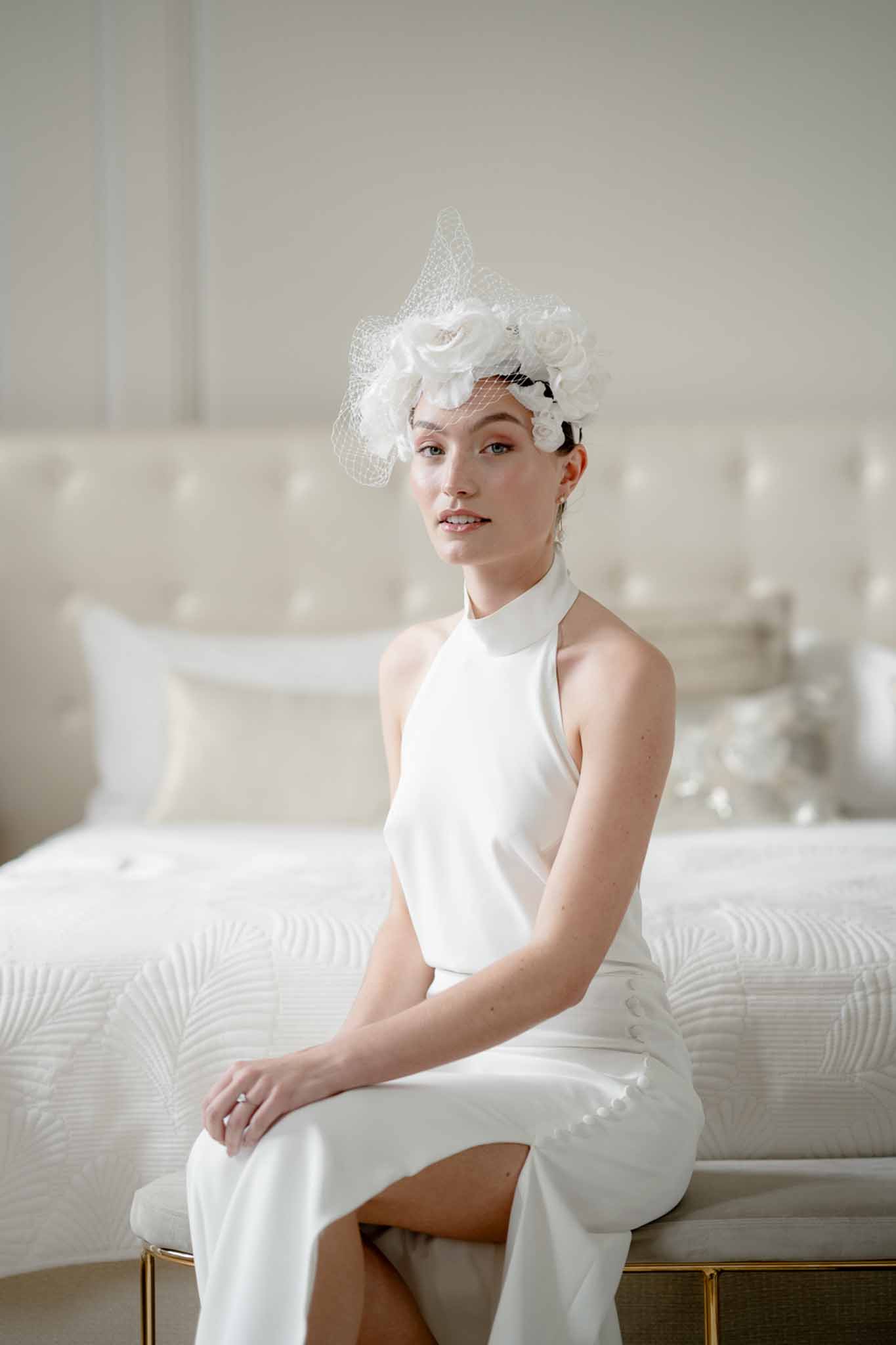 Bride seated on gold bench wearing white halter dress with birdcage fascinator and fabric roses
