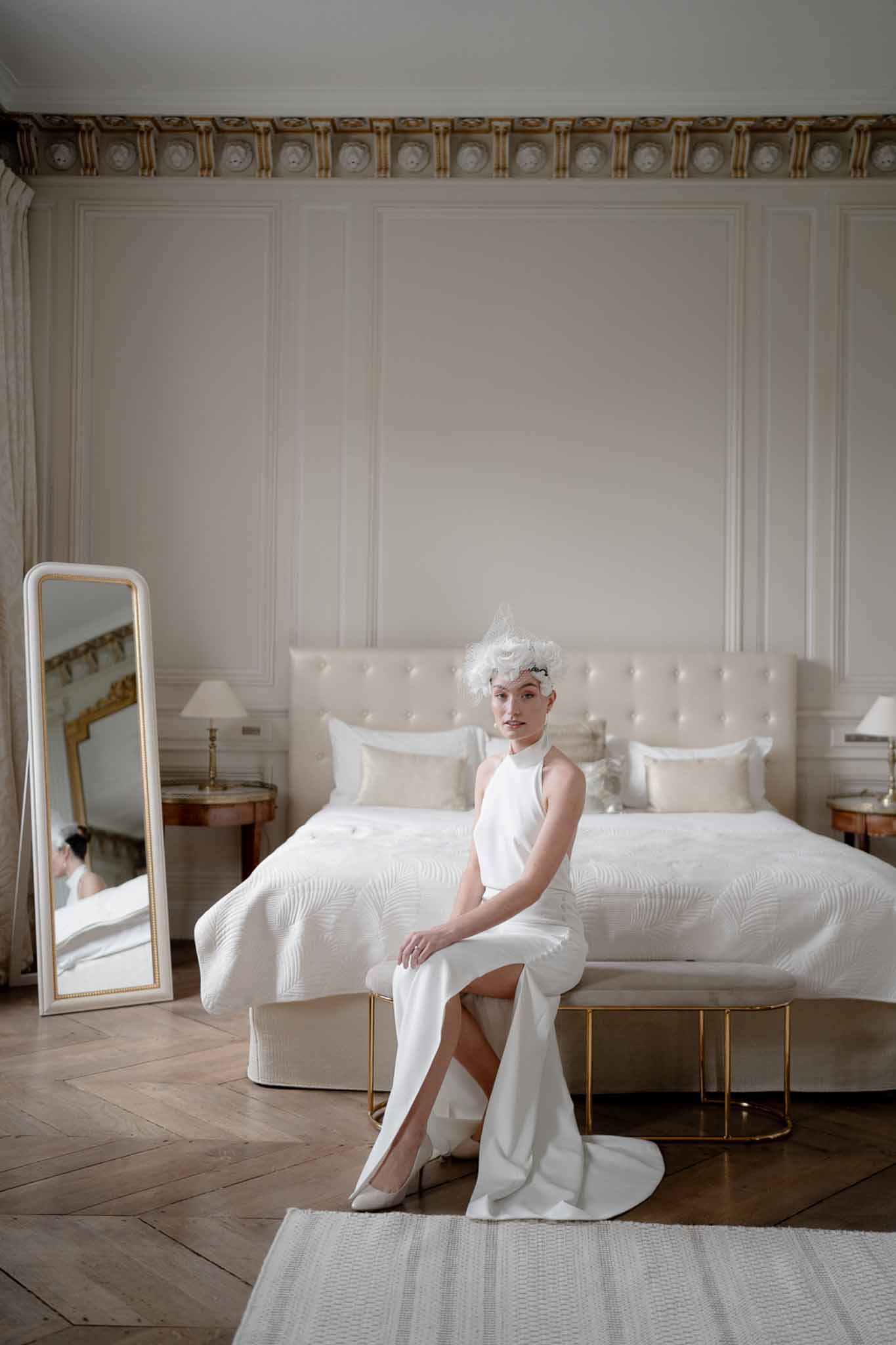Bride in white halter gown with feathered fascinator seated at foot of bed in white boiserie and blush chateau room