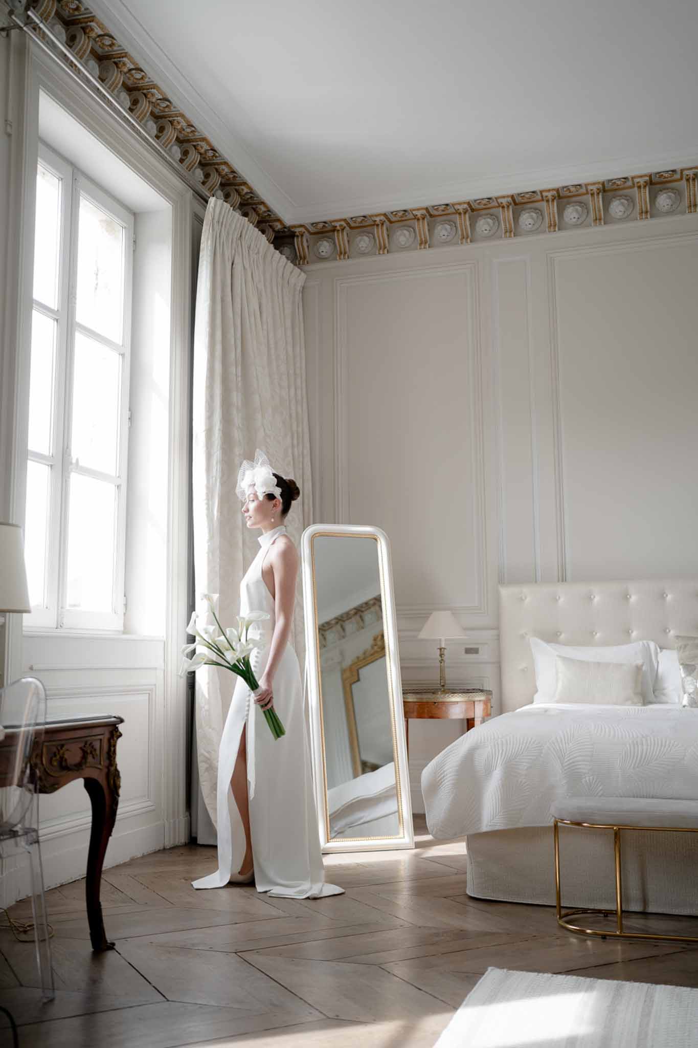 Bride in ivory halter gown with tulle fascinator and calla lily bouquet in gold-accented chateau bedroom