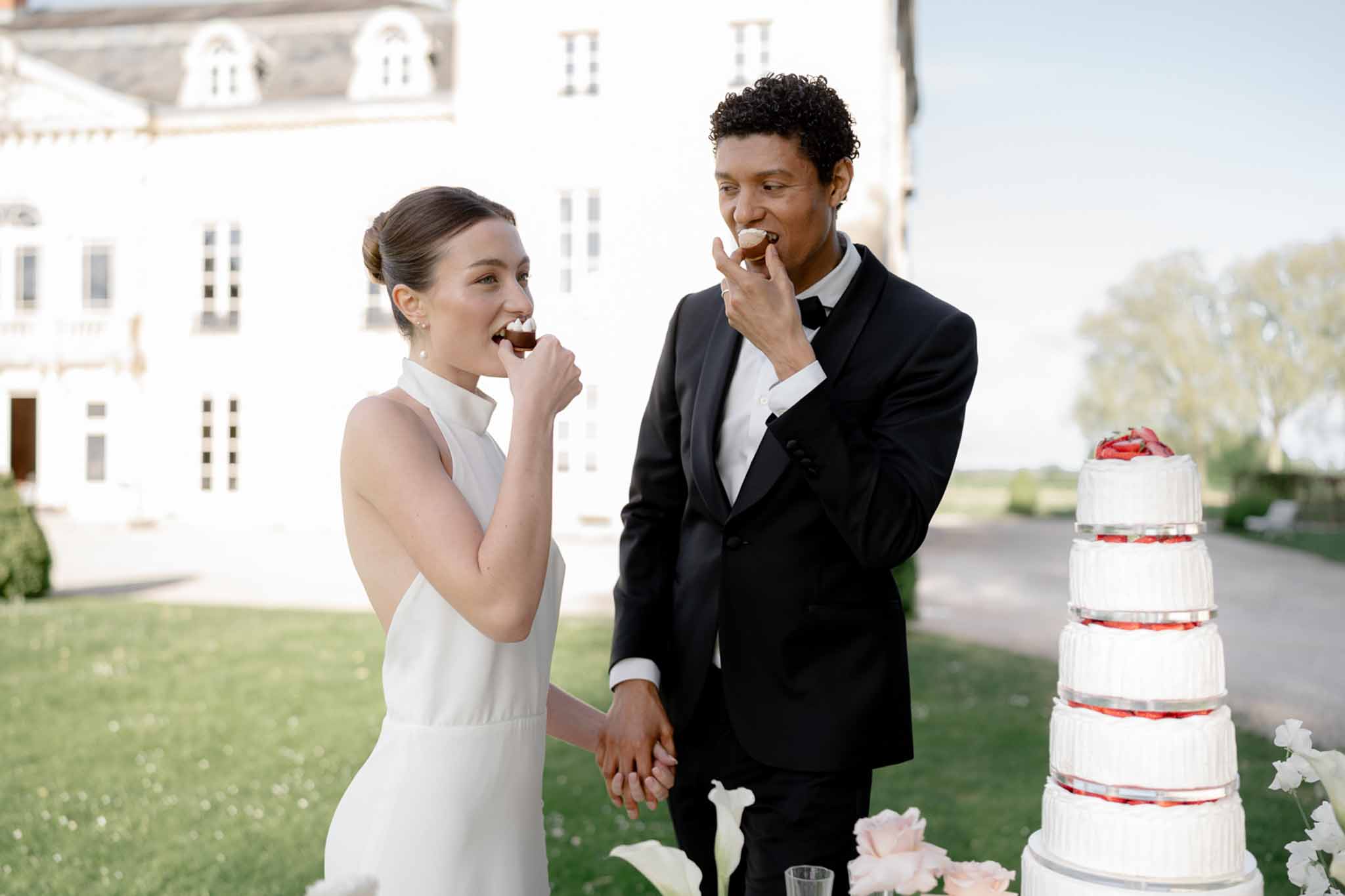 Bride and groom sharing first bites of four-tier strawberry wedding cake on chateau grounds in evening light
