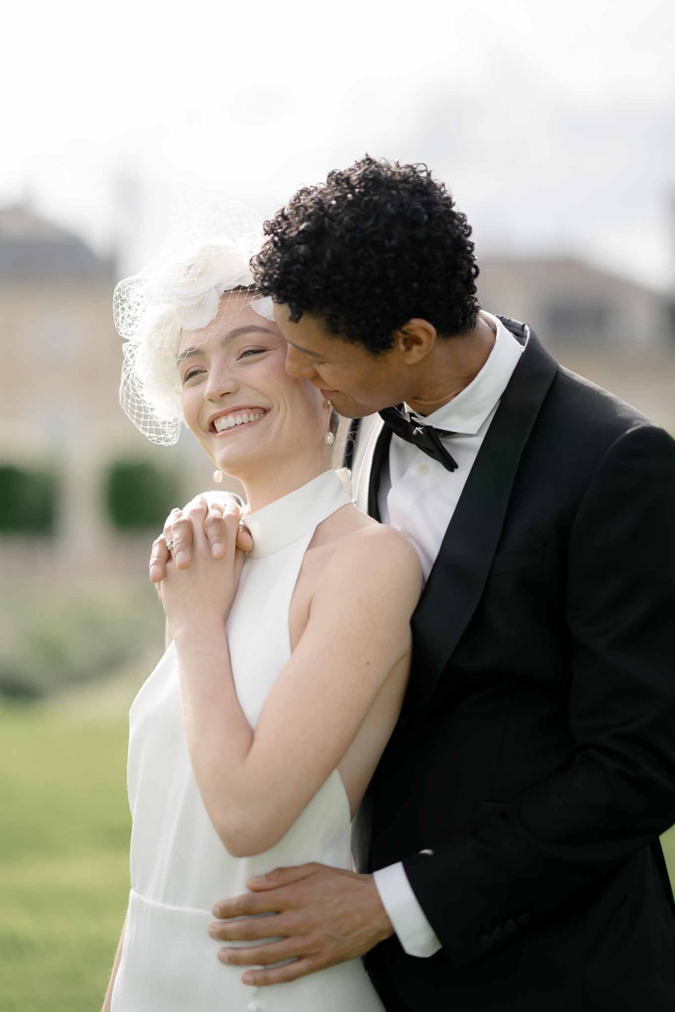 Bride in white halter-neck dress with birdcage fascinator laughing as groom in black tuxedo leans close to her cheek