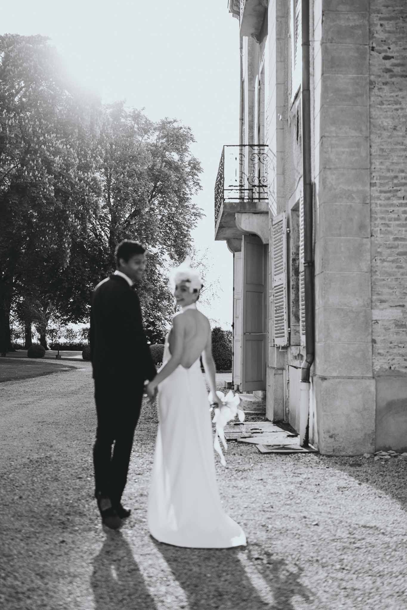 Bride in open-back gown turning back toward camera as groom leads along chateau facade in B&W