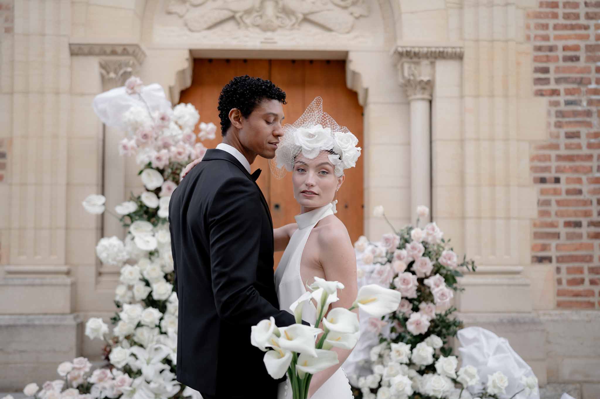 Couple at church entrance with fabric rose headpiece and calla lily bouquet flanked by tall rose arrangements