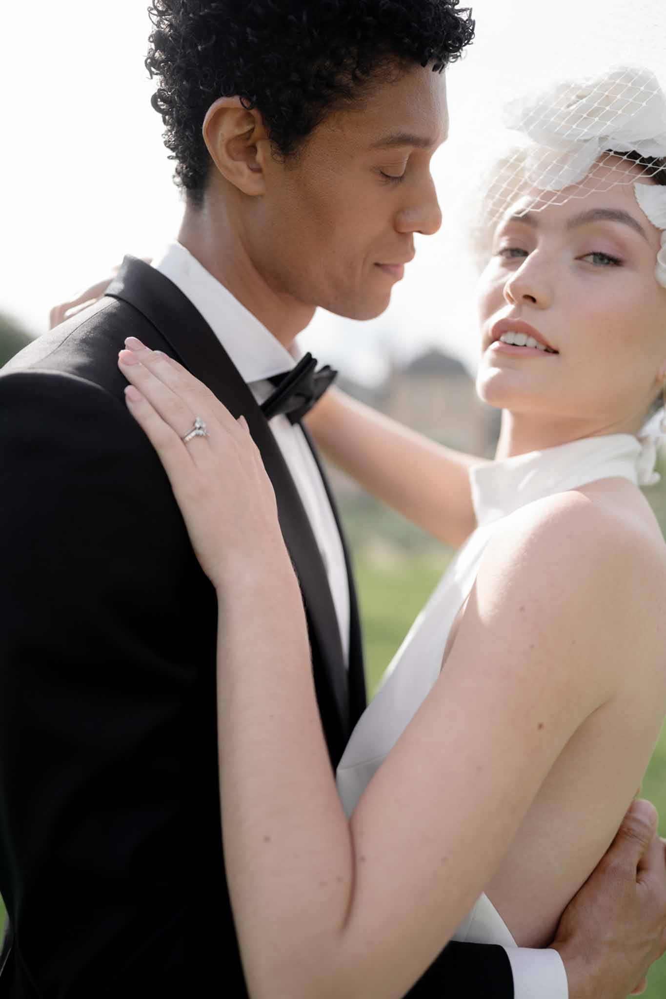 Close-up of bride in birdcage veil and groom in black tuxedo in intimate near-kiss pose outdoors